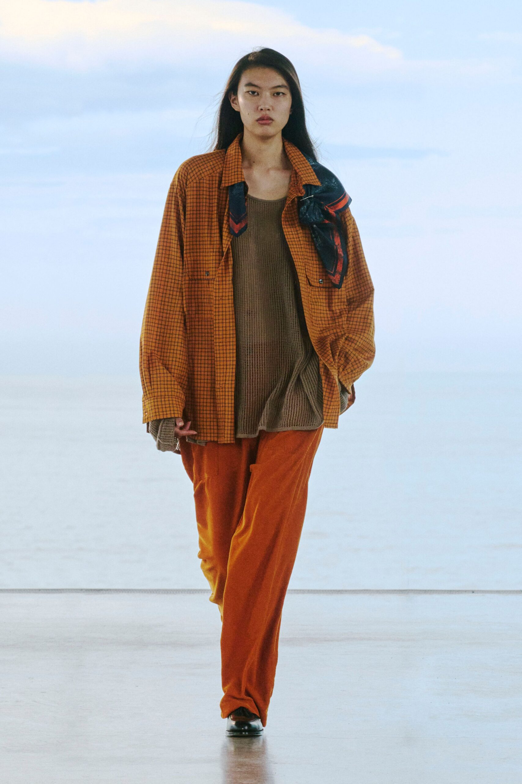 Oversized rust micro-check shirt with flap pockets, a black-and-red scarf pinned at one shoulder. Loose taupe mesh tank with a rolled hem, cinnamon velour trousers, black square-toe shoes. Airport tram at dusk.