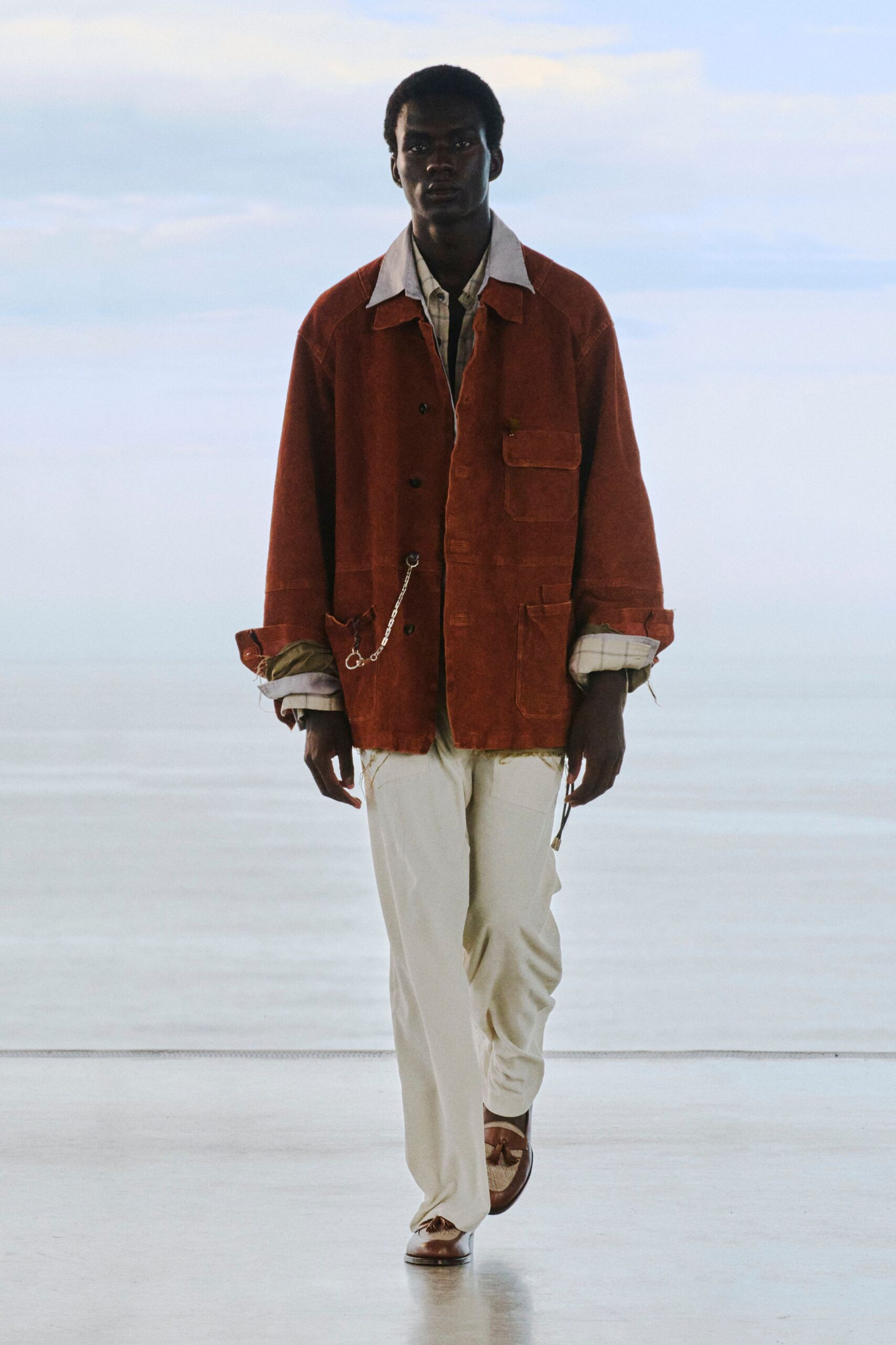 Rust suede chore jacket with patch pockets and a wallet chain clipped at the hip, over a pale checked shirt with turned-back cuffs. Cream drawstring trousers, brown loafers with tasseled vamps. Waiting out wind on a ferry deck.