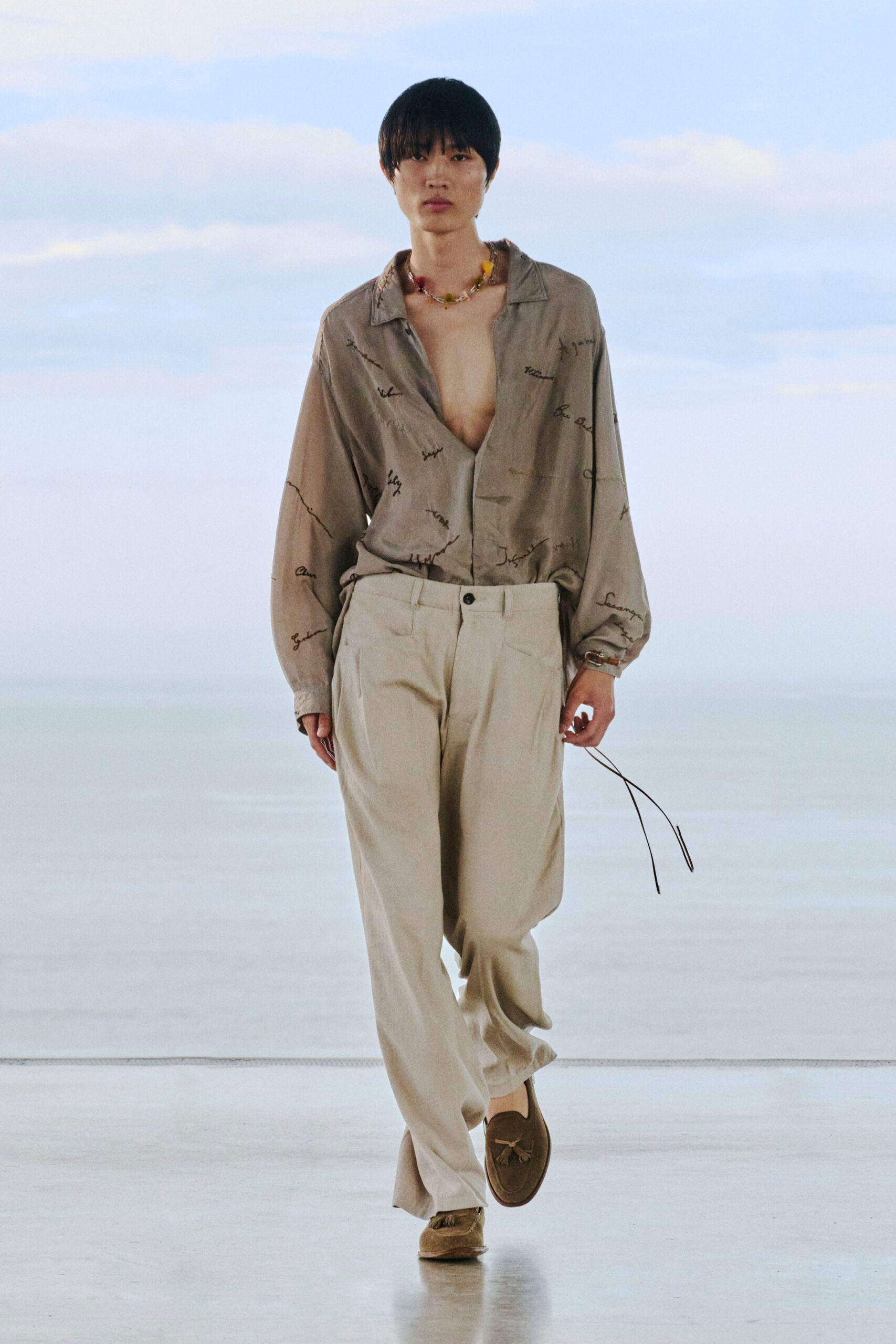 Taupe satin shirt scattered with cursive embroidery, worn open at the chest. Cream pleated trousers with a soft break over brown suede loafers, tassels brushing the toe. A fruit-bead necklace. Late checkout, sea fog still on the windows.