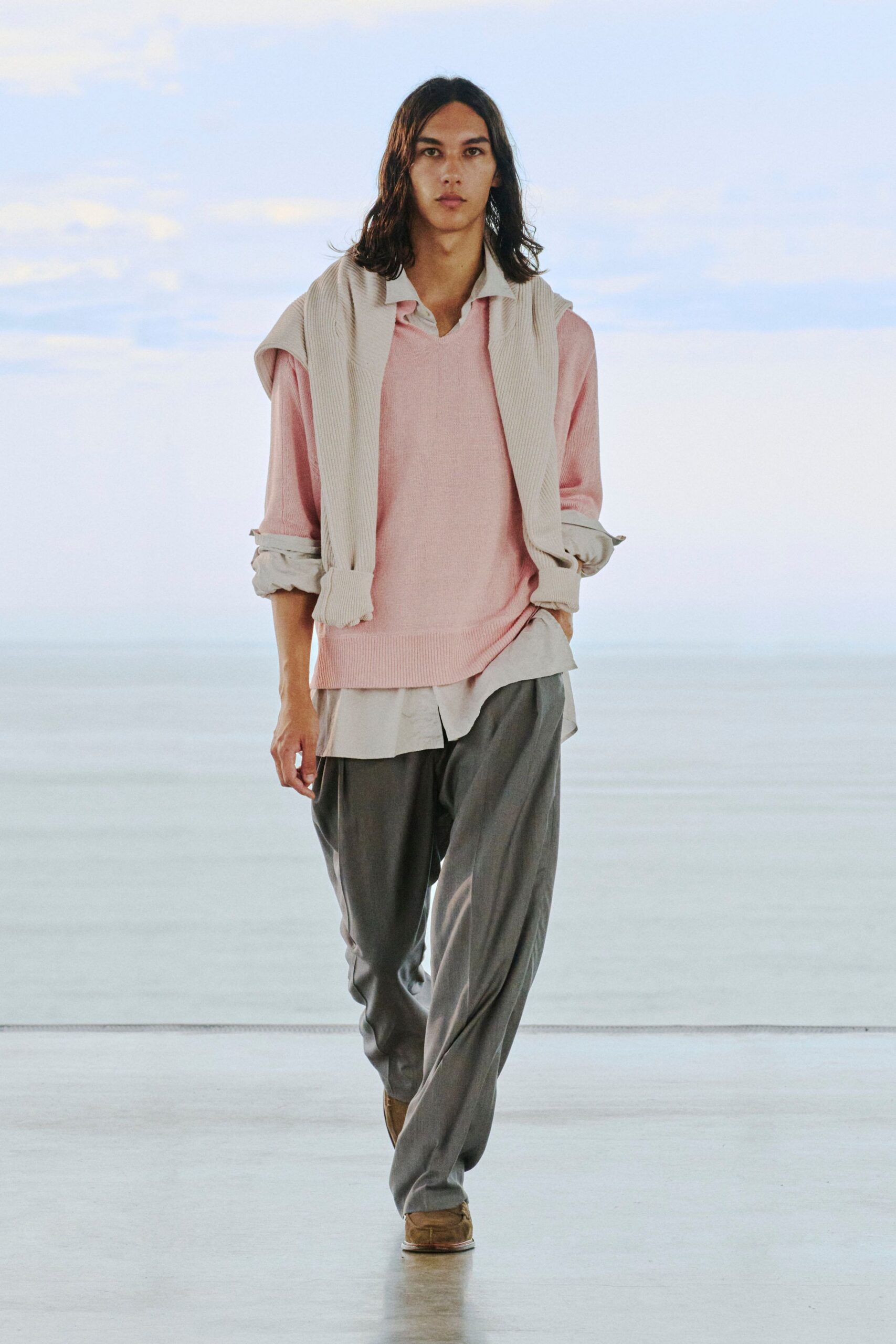Cream ribbed knit draped over the shoulders, pale pink V-neck sweater with a deep waistband, chalk button-down with the shirttail slipping below the hem, grey trousers with a soft break, tan suede shoes. Early ferry, wind still cool off the water.