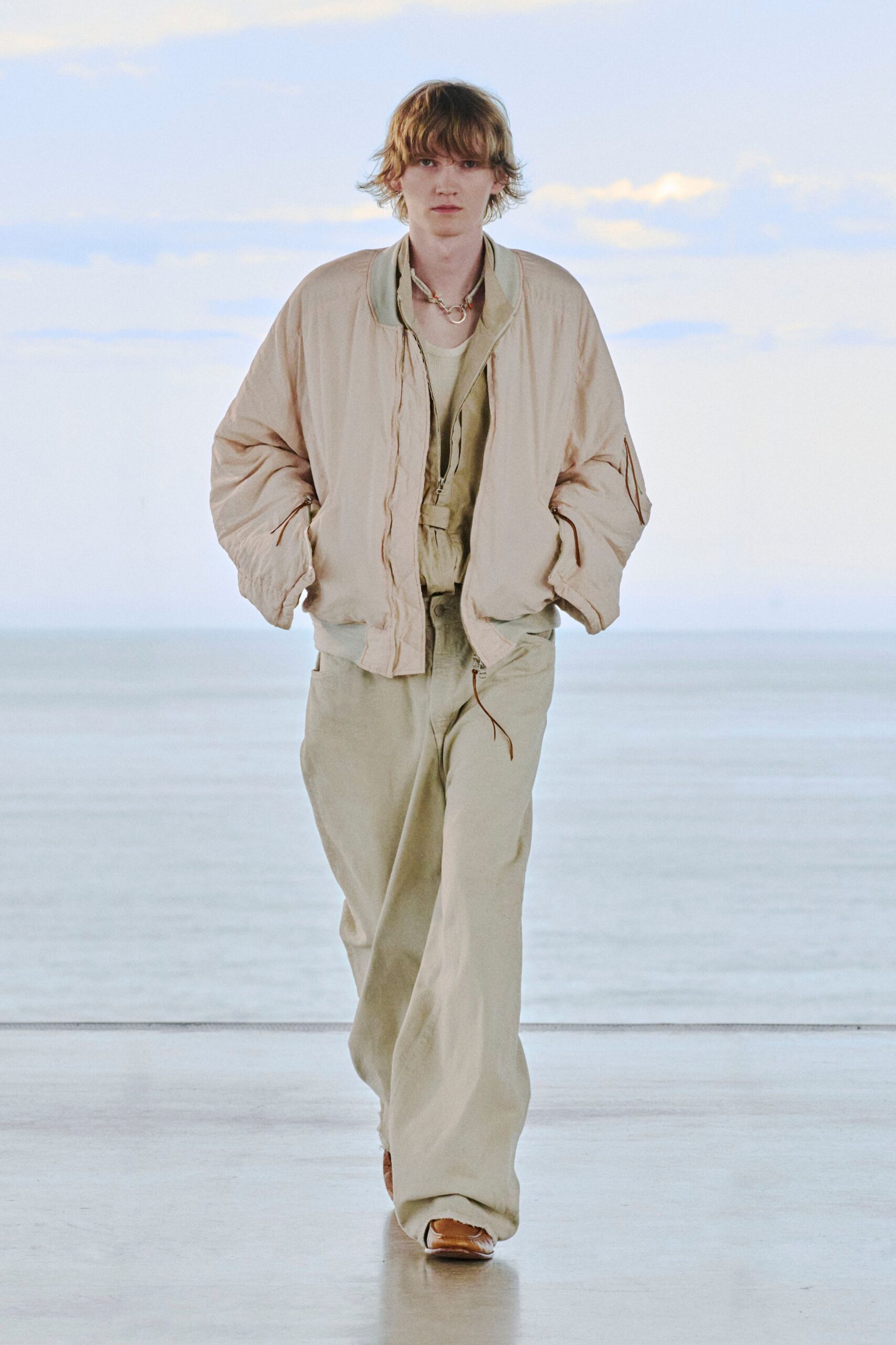 Blush satin bomber with a crinkled sheen and zip pocket at the sleeve, over a pale ribbed tank and washed sage wide-leg trousers with a dangling zip tape. Tan square-toe shoes. Early ferry, salt still on the rail.