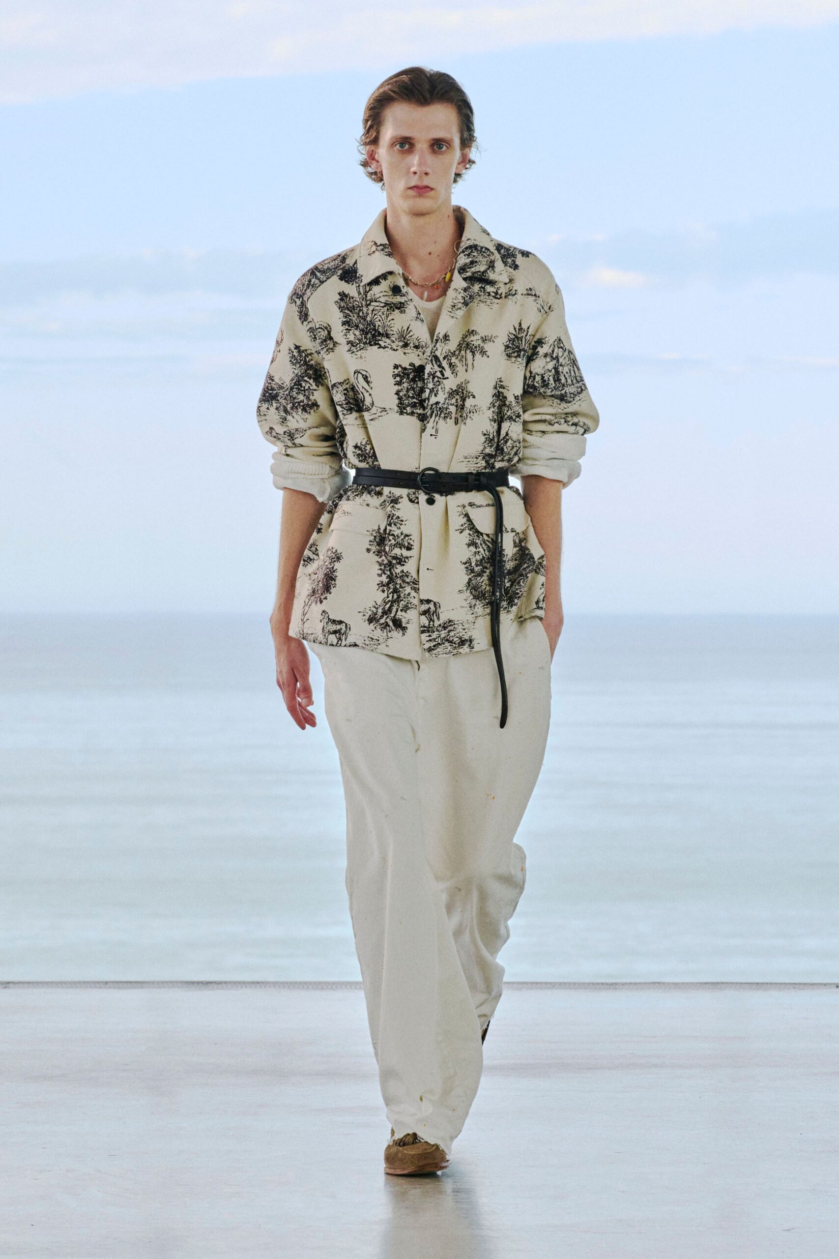 Cream toile jacket printed with black pastoral scenes, cinched by a skinny leather belt with the tail left hanging. White ribbed crewneck underneath, chalky loose trousers, tan suede shoes. Early ferry, wind off the water.