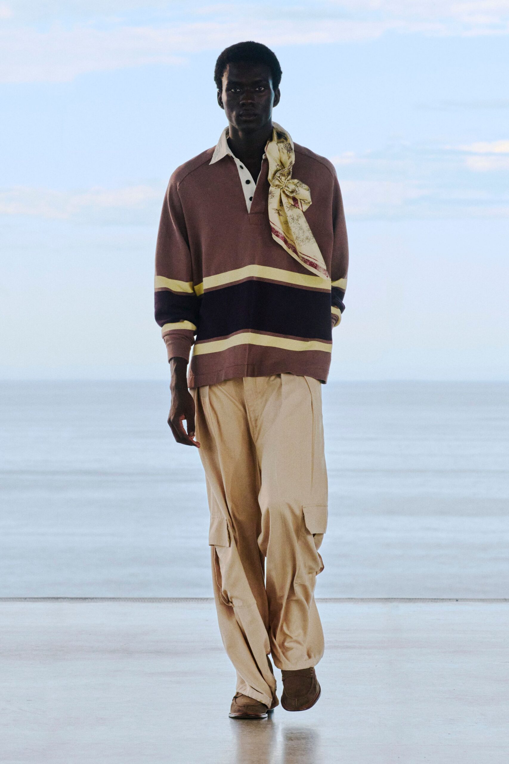 Striped rugby shirt in dusty mauve knit with a white placket and black buttons, cream printed silk scarf knotted at one shoulder, sand cargo trousers with flap pockets, brown suede shoes. Early ferry deck, wind catching the scarf.