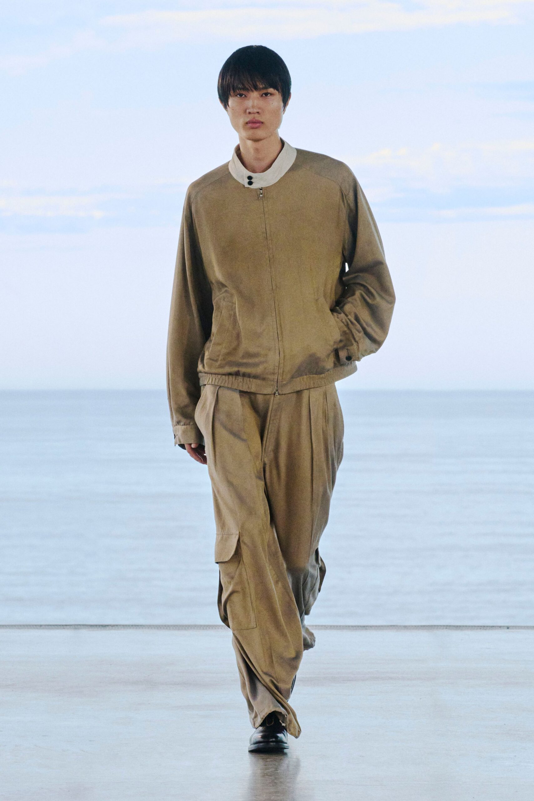 Taupe zip-front blouson in brushed twill, finished with a white stand collar fastened by two black snaps. Loose sand cargo trousers with deep knee pleats, black square-toe shoes. Early ferry, salt still on the rail.