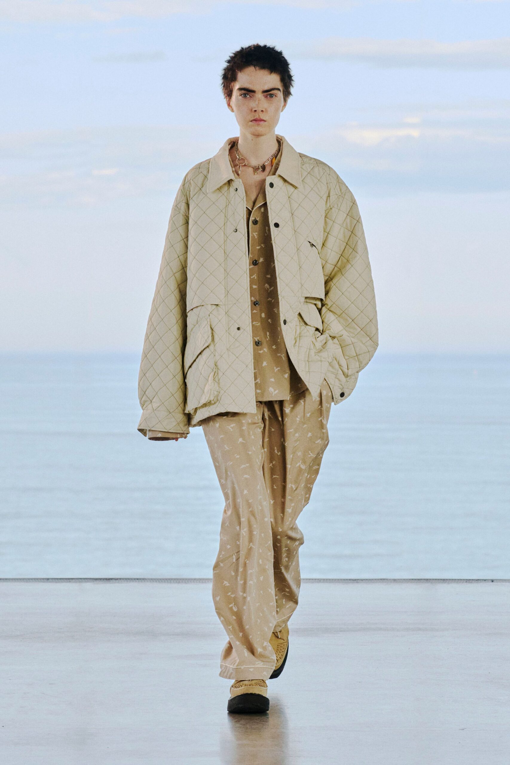 Cream diamond-quilted field jacket with flap pockets and a small zip welt at the chest, over a sand shirt scattered with tiny white marks. Matching loose trousers, woven slip-ons. Ferry deck at first light, salt still in the air.