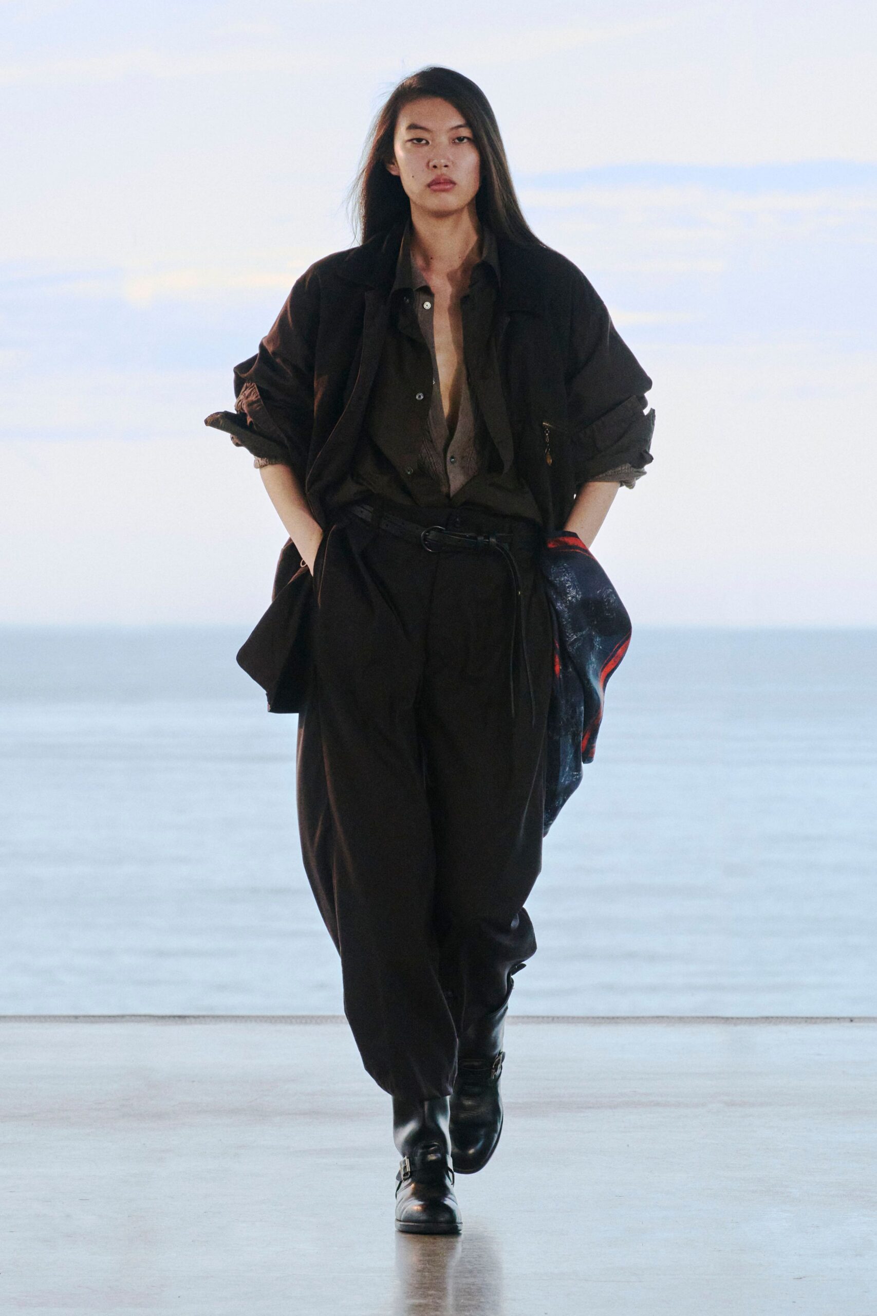 Slouchy black jacket with rolled cuffs over a dark olive shirt hanging open on mismatched buttons. High-waisted black trousers cinched with a skinny belt, square-toe harness boots. Dawn ferry deck, salt in the air.