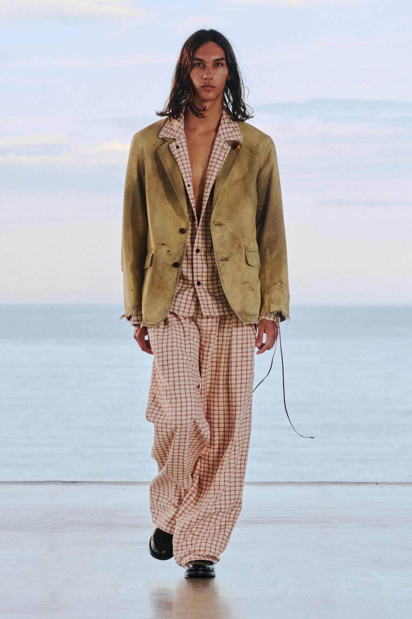 Olive suede blazer with frayed cuffs and little scuffs, worn over a cream shirt printed in red windowpane checks. Matching loose checked trousers puddle over black square-toe shoes. Sea air at dusk, waiting out a missed ferry.