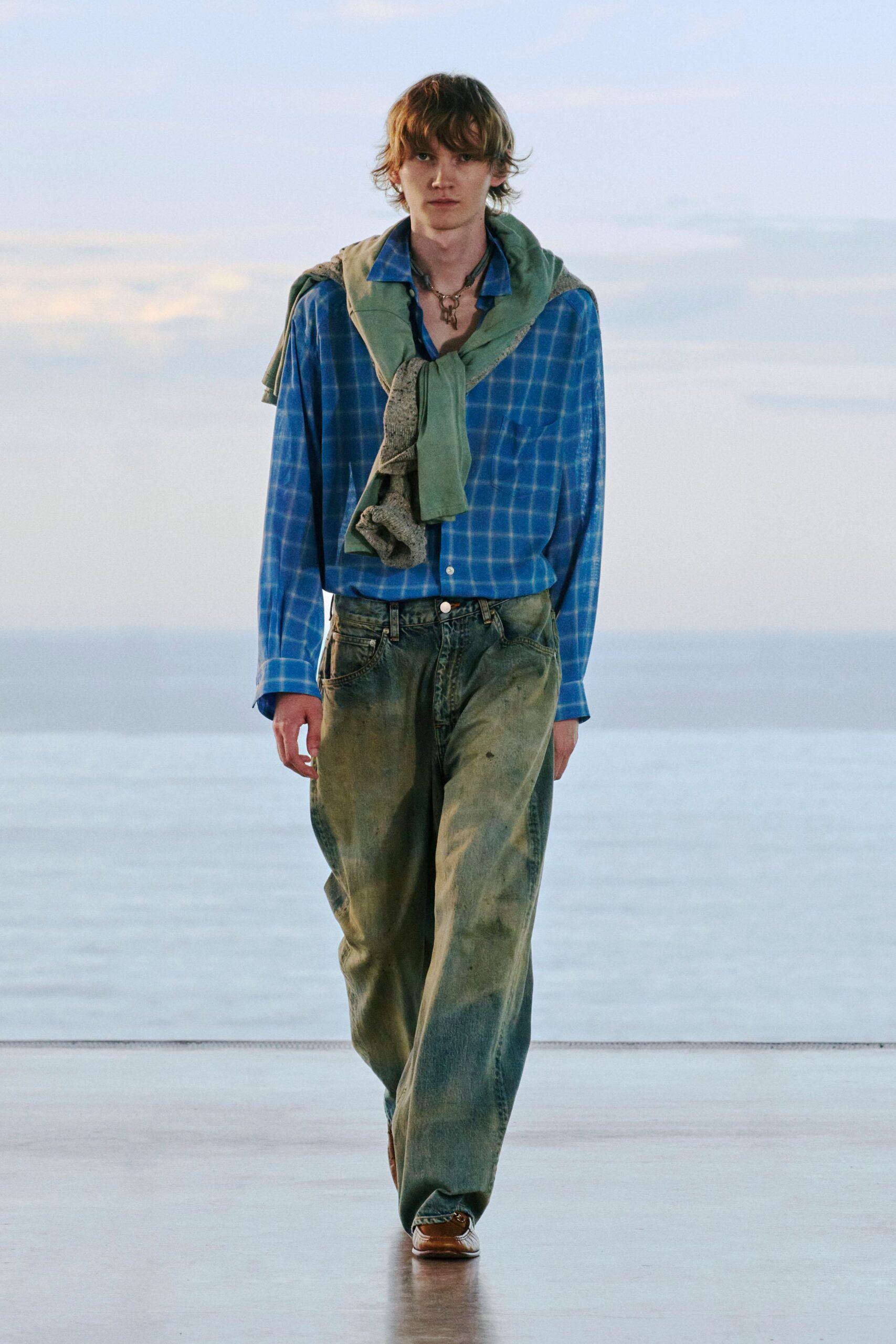 Blue windowpane shirt with a soft sheen, collar left open. Sage sweatshirt draped and knotted at the chest, speckled knit peeking from the underside. Faded wide-leg jeans, tan square-toe shoes. Cold boardwalk at first light.