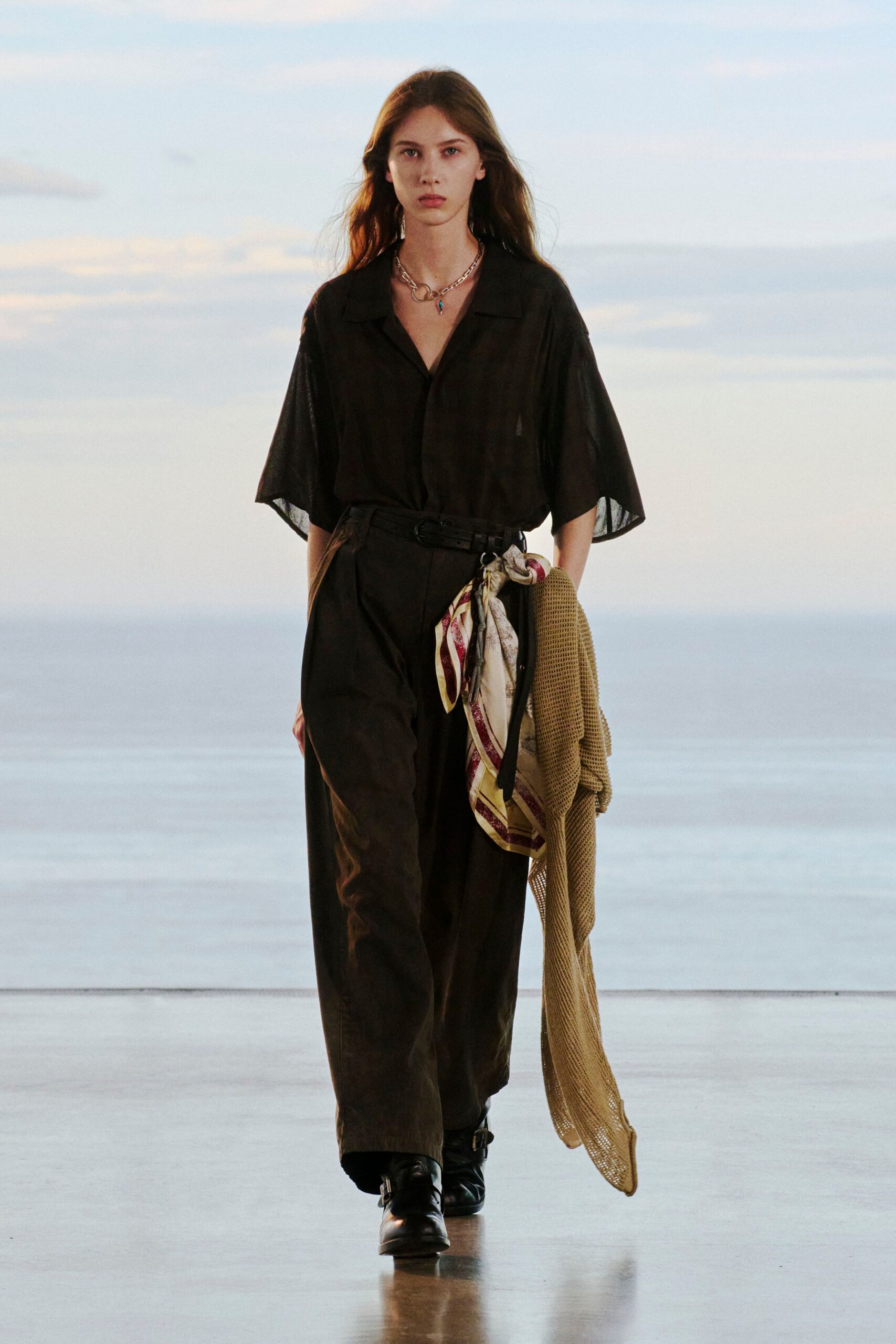 Black gauze camp-collar shirt with airy elbow sleeves, loose black trousers cinched by a narrow belt with a striped silk scarf knotted through the ring. Black buckle boots, sand netting in hand. Dusk on a ferry deck.