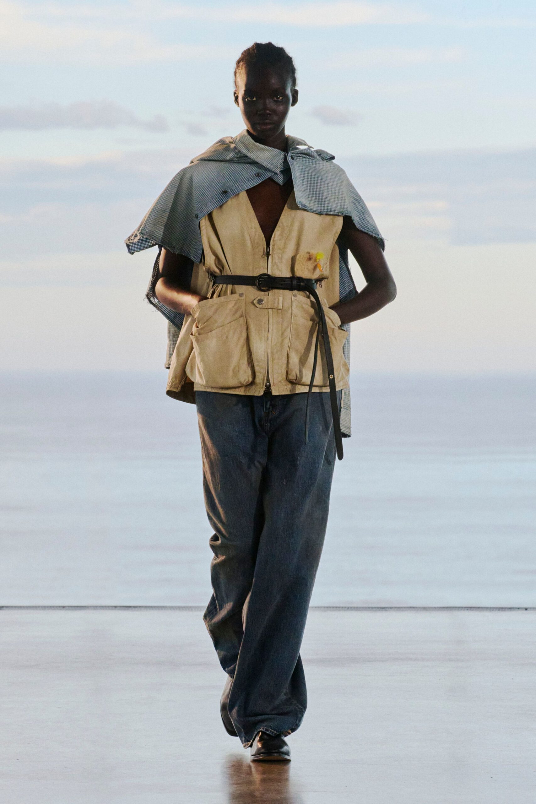 Blue micro-check cape with raw-cut edges draped over a sand utility vest, zipped low and loaded with bellows pockets. Narrow black belt hanging long, washed indigo jeans, square-toe shoes. Wind off the pier at dusk.