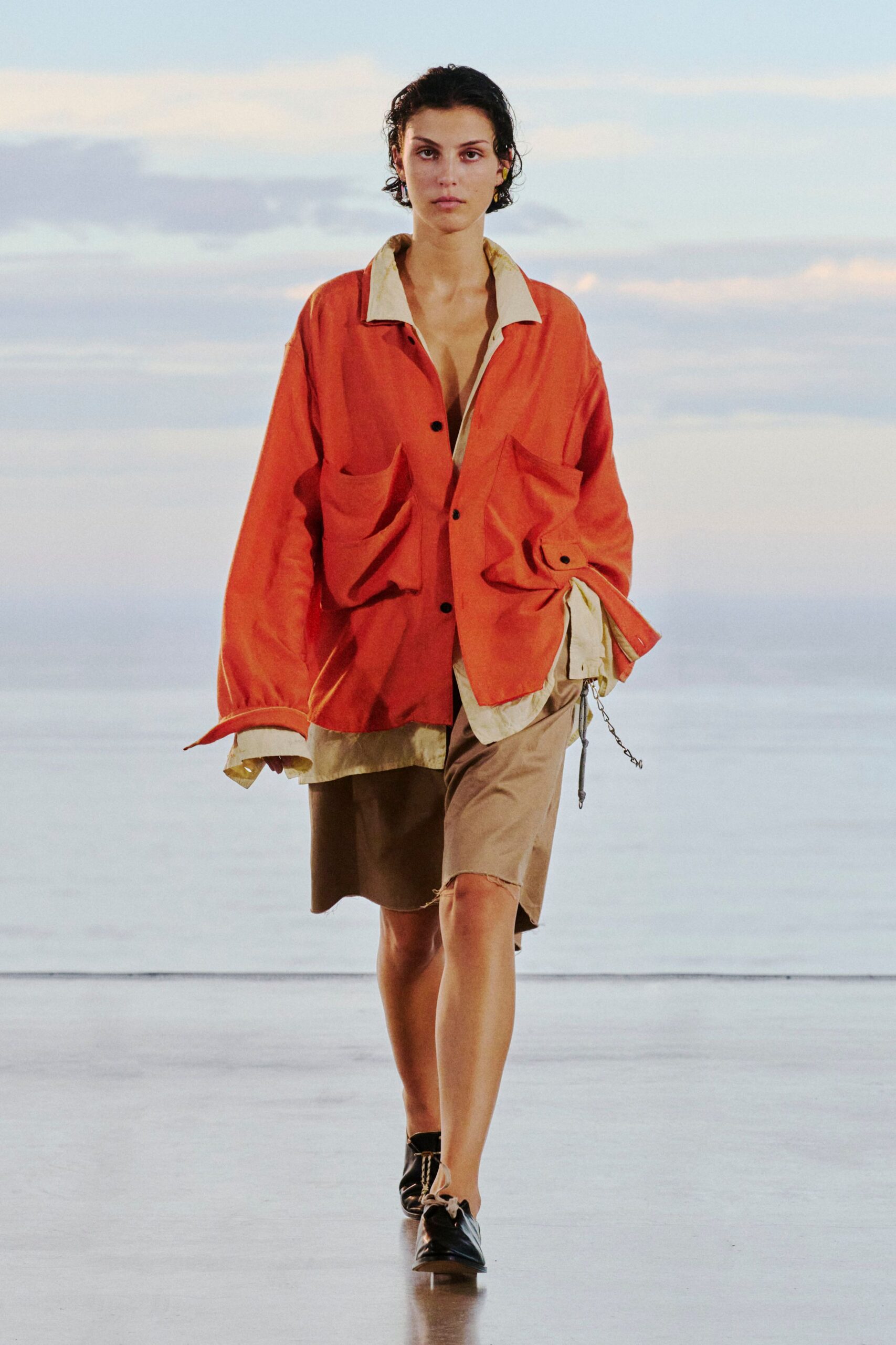 Persimmon utility jacket with oversized bellows pockets and loose cuff tabs over a cream shirt in washed poplin. Taupe knee-length shorts with a raw-cut hem, black lace-up flats. Early ferry, salt drying on your shins.