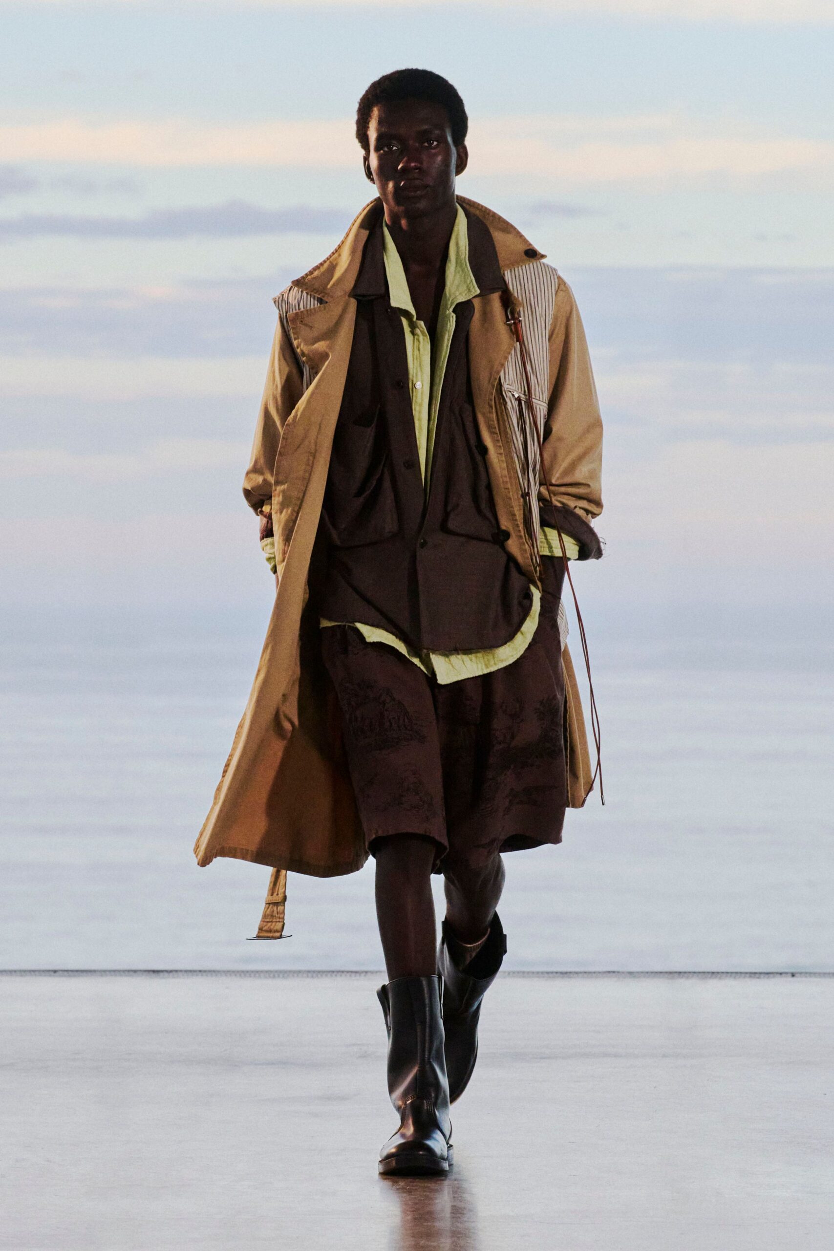 Sand trench coat with a striped inset panel and a strap end swinging at the hem, over a dark camp-collar shirt and crinkled celadon layer. Dark shorts with tonal toile, black ankle boots. Dusk on a ferry deck, salt in the air.