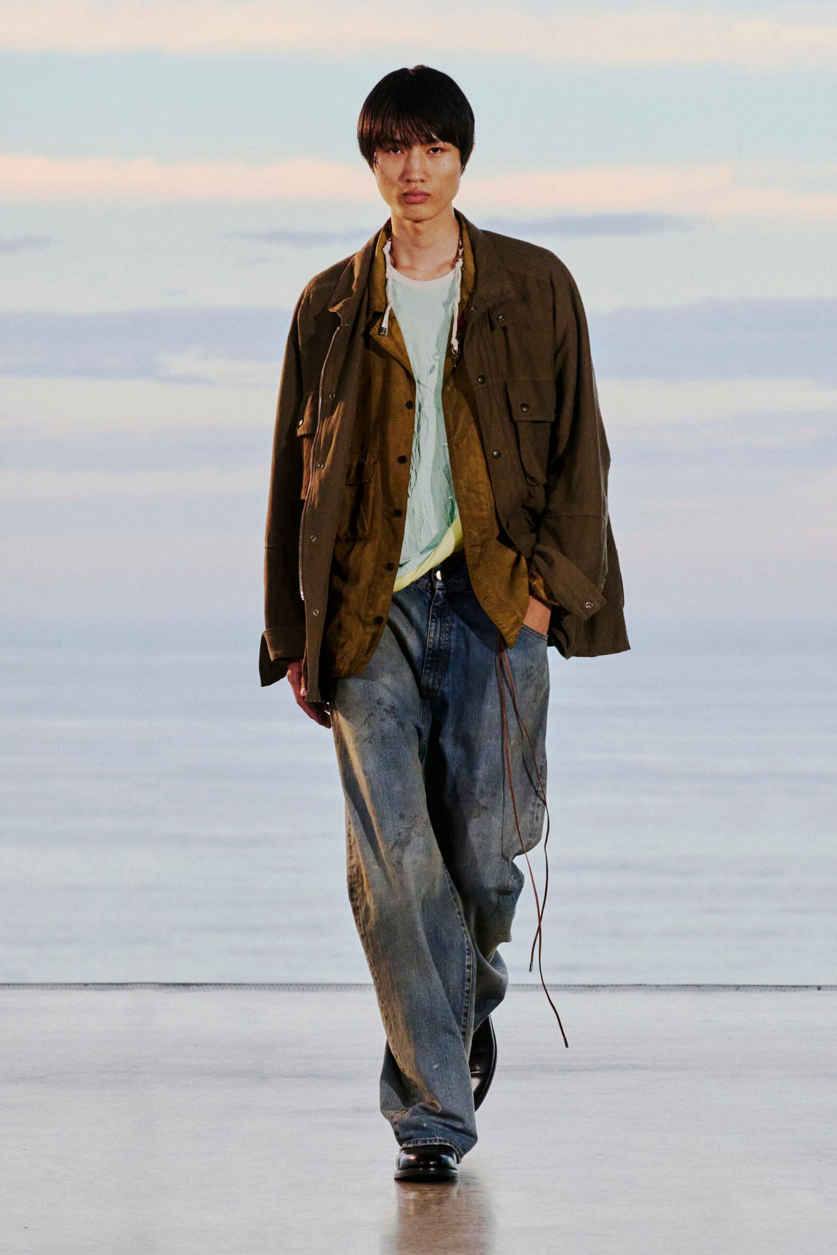 Olive field jacket with snap-flap pockets over a bronzy crinkled overshirt, pale mint tee, and washed blue jeans slouched over square-toe black shoes, one leather drawcord trailing low. Cold air off the water at dusk.