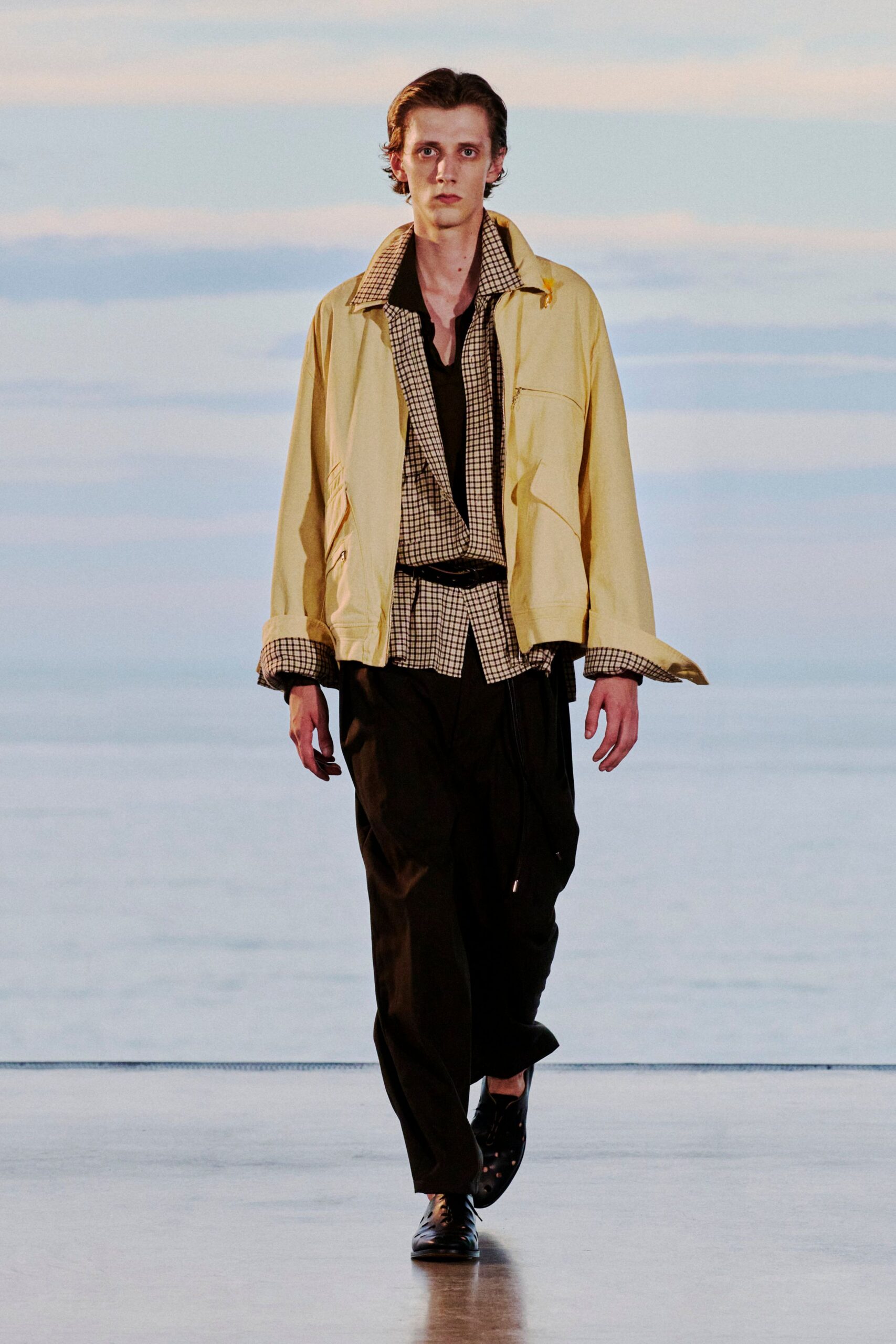 Butter-yellow zip jacket with checked facing turned out at the collar and cuffs, over a small-grid shirt cinched by a skinny black belt, black draped trousers, punched leather derbies. Waiting for the last ferry in a wind off the water.