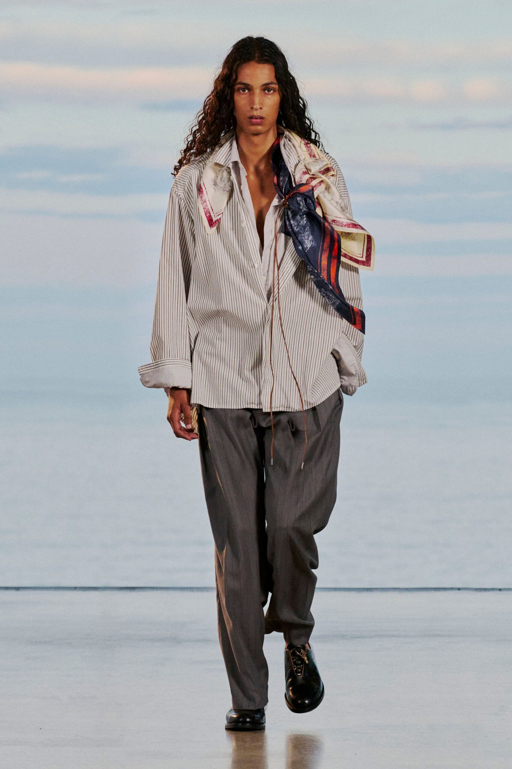 Oversized banker-stripe shirt in crisp poplin, cuffs turned back to show the white facing. Silk scarf knotted off one shoulder, charcoal trousers with a loose break, black leather derbies. Ferry deck at dusk, salt in the air.