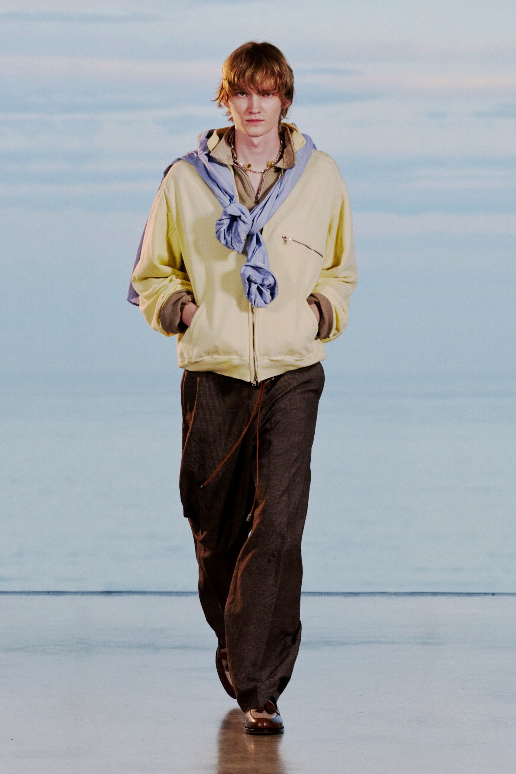 Pale lemon zip-front jacket with a slanted chest zip, taupe hooded top peeking out beneath, powder-blue shirt knotted around the shoulders, dark brown drawstring trousers, square-toe leather shoes. Seaside hotel corridor at first light.