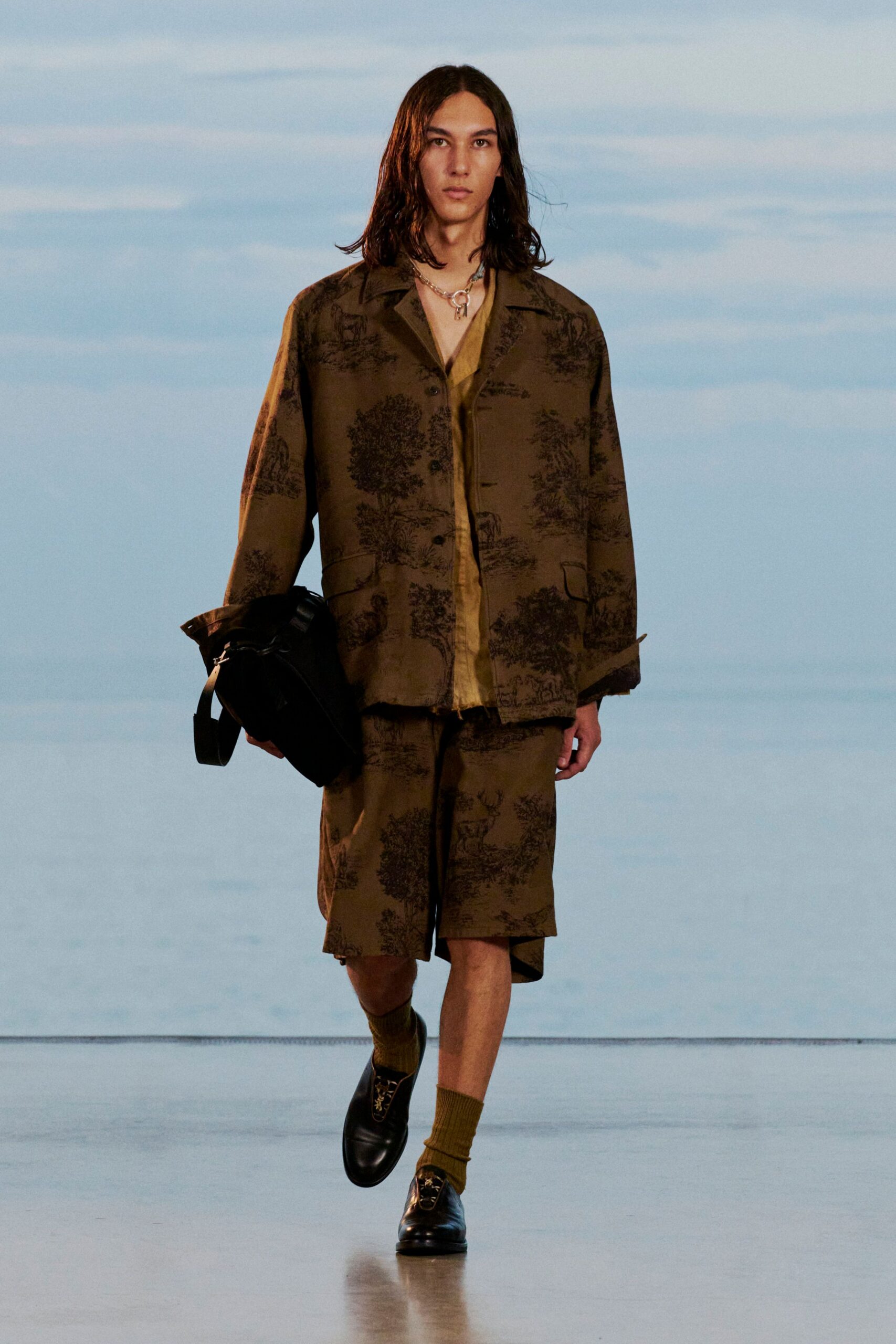 Brown camp-collar jacket printed with pastoral toile, over a honey silk shirt left open at the throat. Matching knee-length shorts, olive ribbed socks, black loafers with horsebit hardware, slouchy black bag. Ferry deck at dusk.