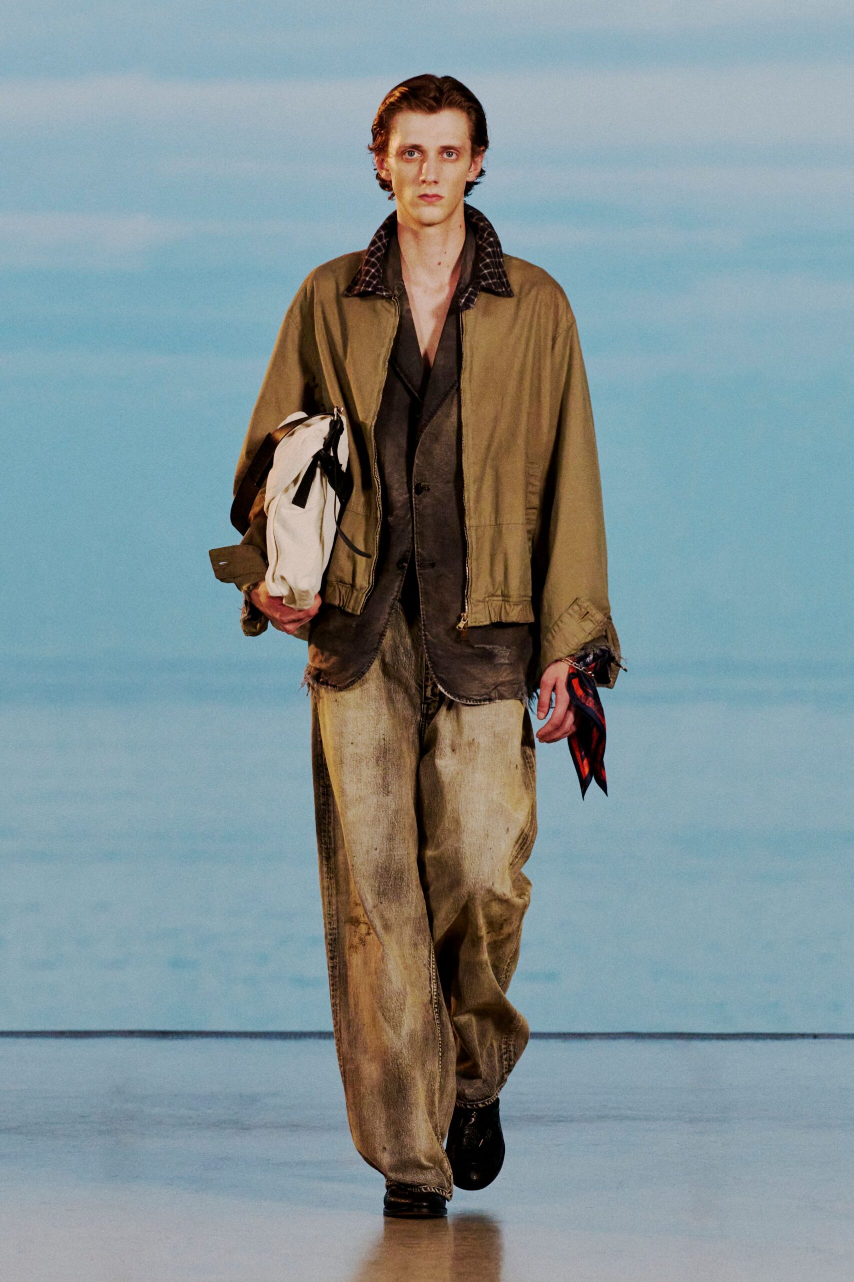 Olive cotton blouson with a dark checked collar and a zip stopped short of the hem, over a washed black jacket with raw edges. Grimed stonewashed jeans, black shoes, white duffel. Dawn at a rural bus depot.