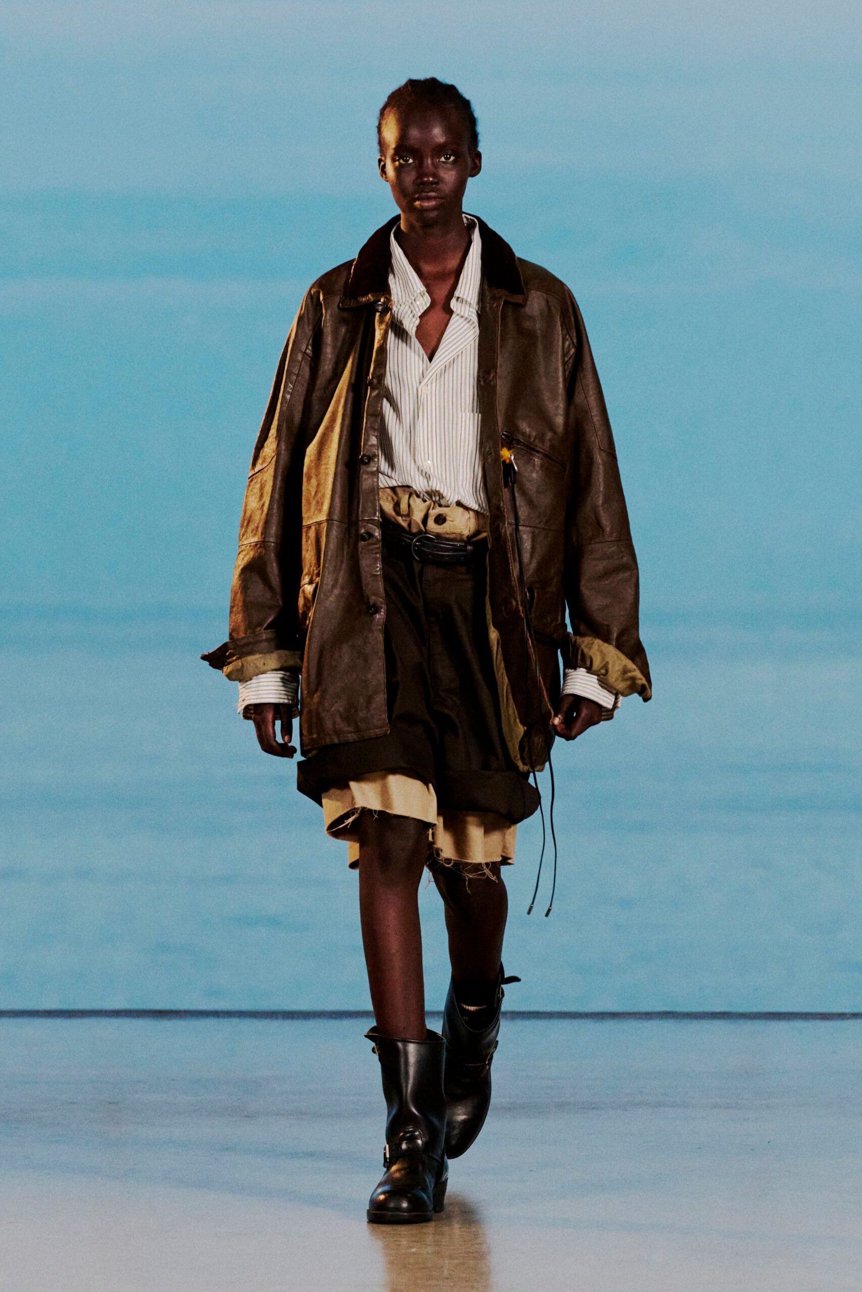 Oversized brown leather jacket with a dark velvet collar, over a pale striped shirt with long cuffs. Belted layered shorts with a frayed beige underlayer, black ankle boots. Dawn at the ferry terminal, coffee gone cold.