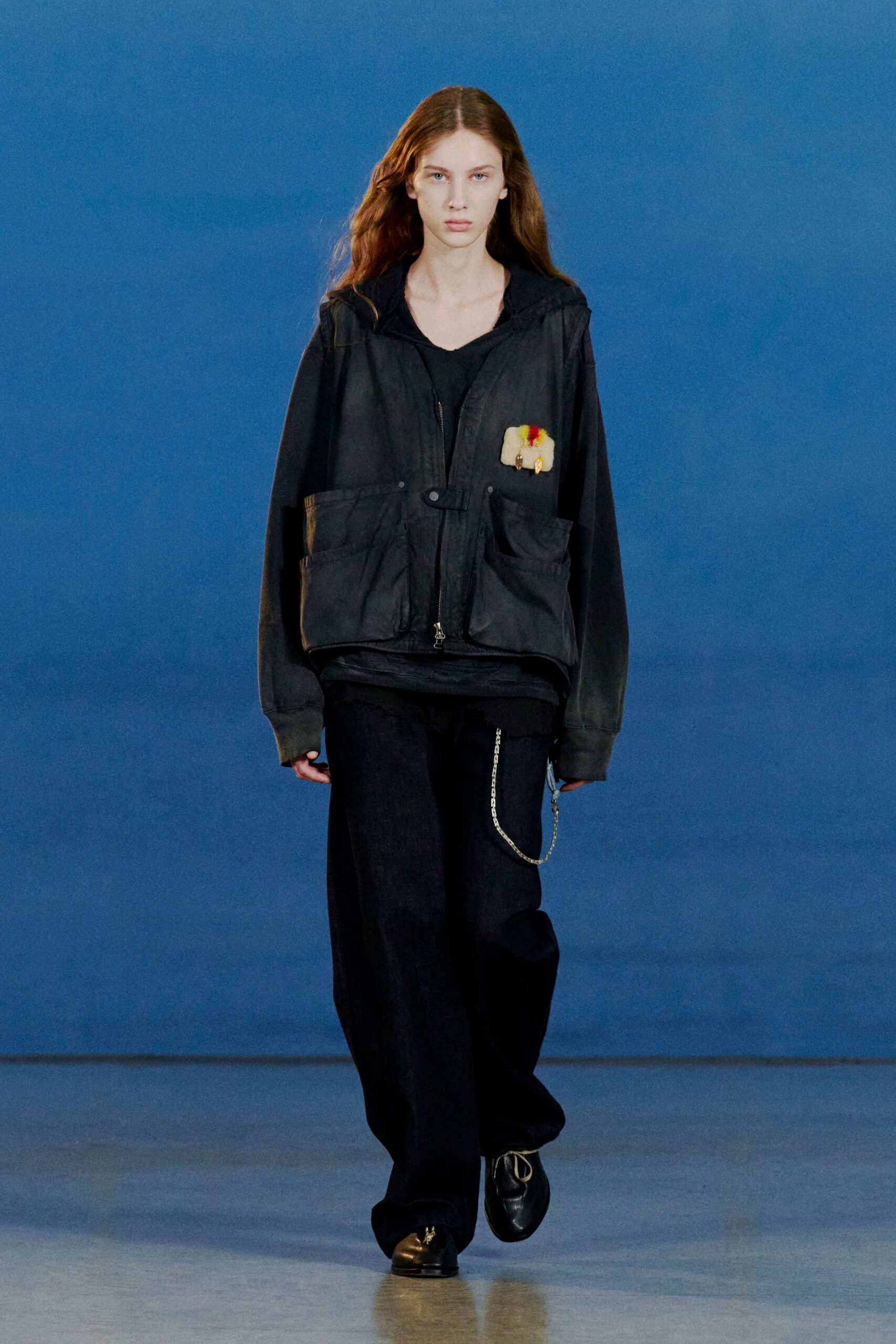 Black hooded jacket in washed cotton, cut with oversized bellows pockets and a fuzzy cream patch at the chest. Black V-neck top, pooled wide-leg trousers with a silver wallet chain, square-toe shoes. Bus depot at dusk, vending machine humming.