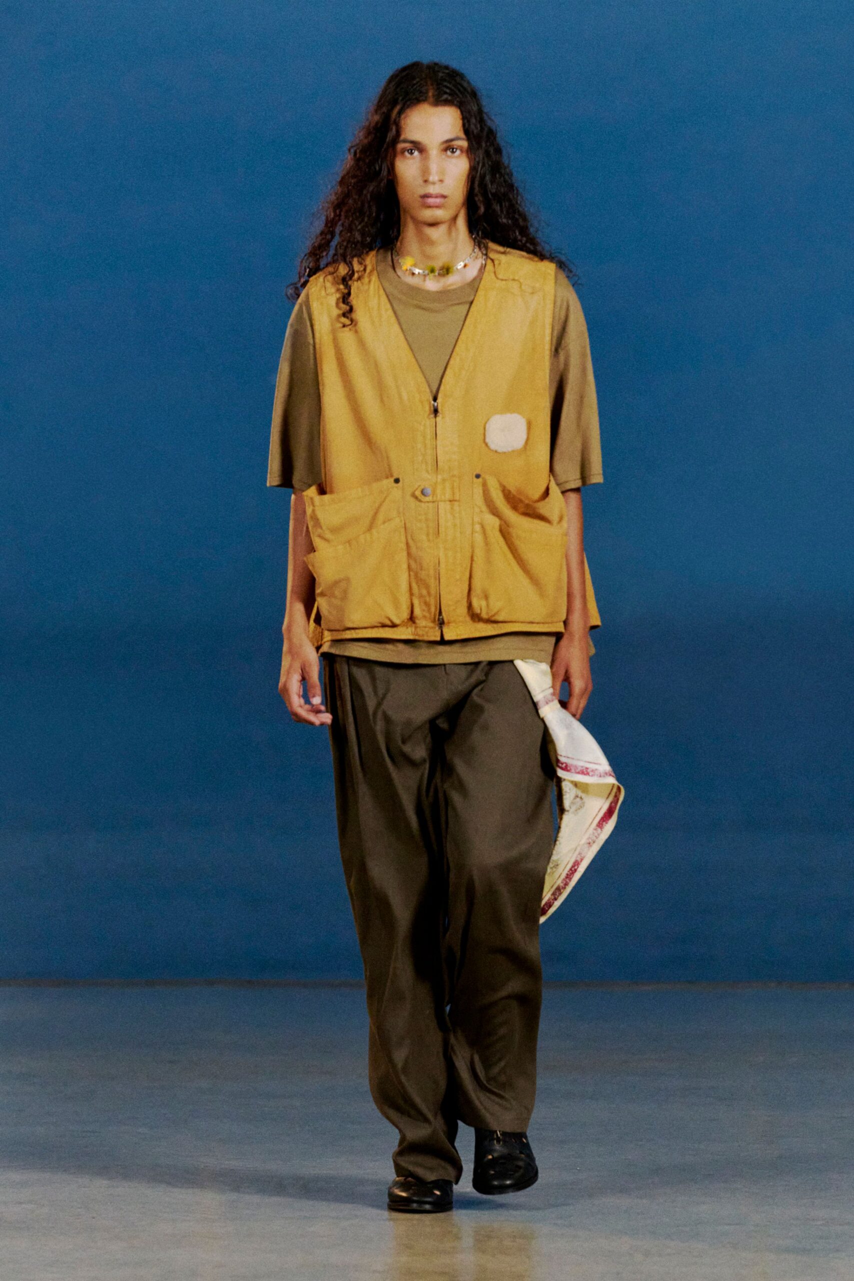 Mustard utility vest in washed canvas, zipped and slouched with bulging patch pockets and a white oval patch; olive crewneck tee with dropped sleeves, loose dark trousers, black shoes. Like heading for the bait shop at dusk.