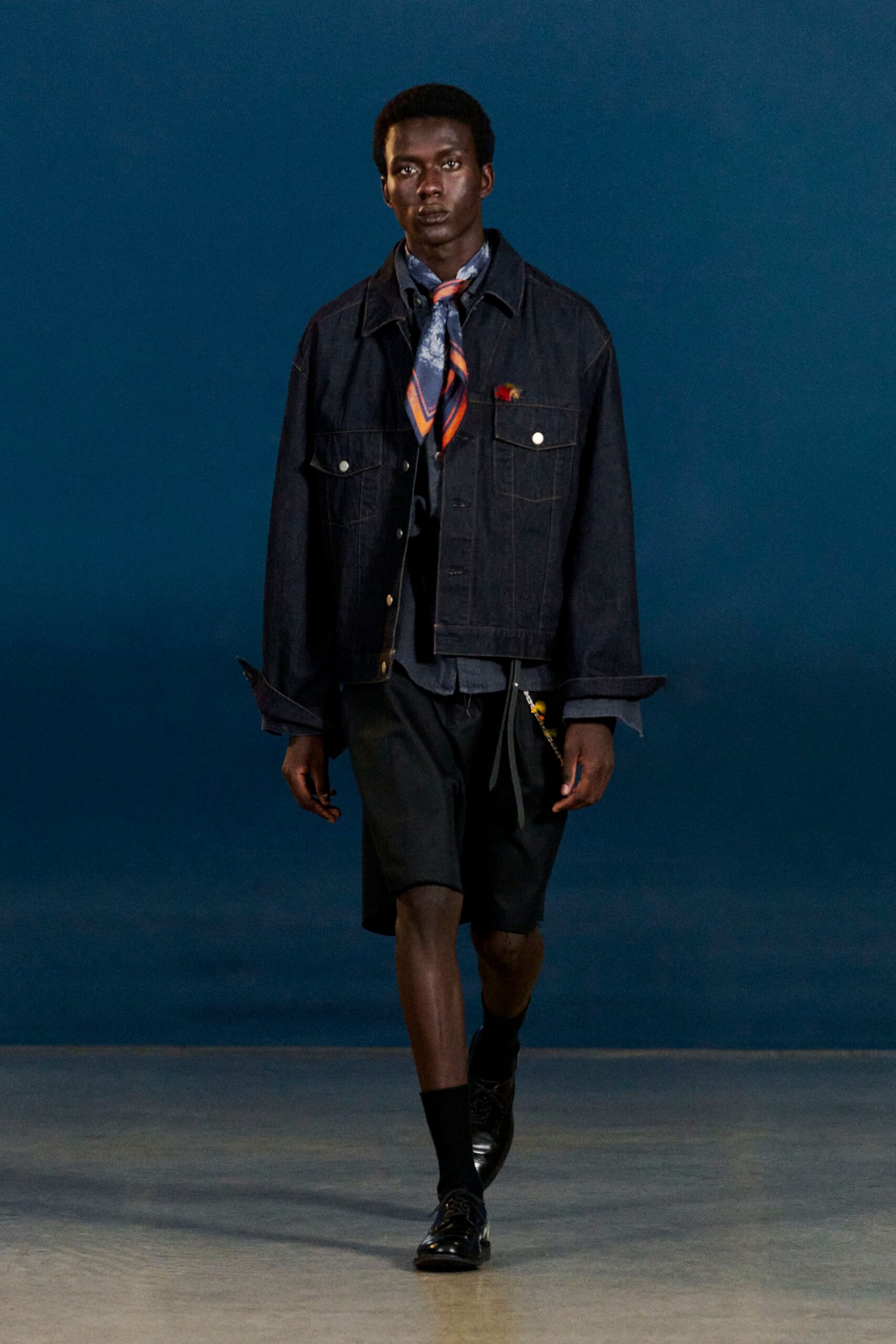 Dark indigo denim jacket with bright metal shank buttons and turned-back cuffs, over a slate shirt and orange-striped silk scarf knotted loose. Black knee shorts, ribbed socks, square-toe derbies. Waiting for the ferry after rain.