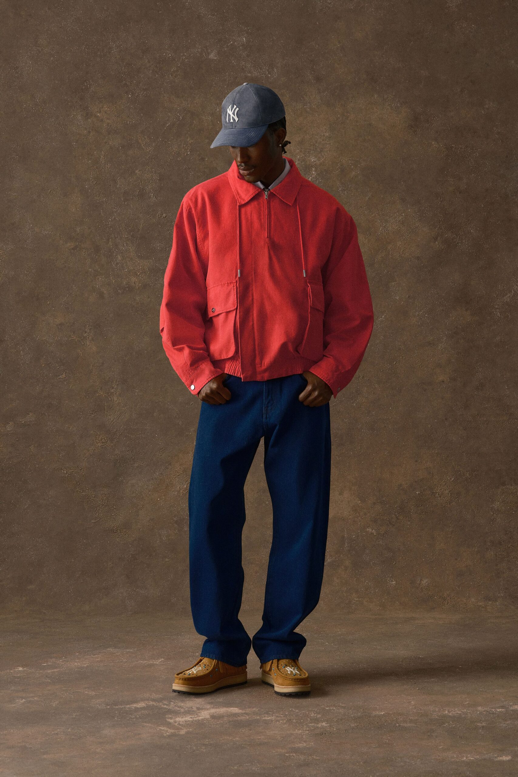 Slate twill baseball cap, coral cotton jacket with a half zip and flap pockets hanging low, pale blue shirt collar peeking out, deep indigo trousers with a loose break, tan suede wallabees stitched with beads. Waiting outside the station at dusk.