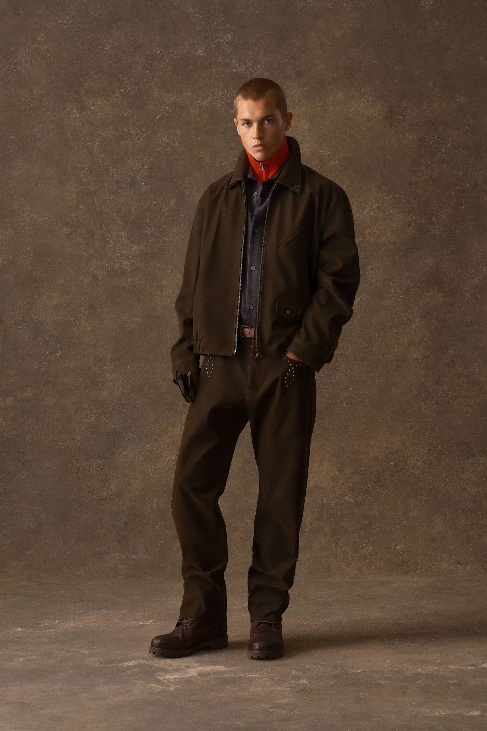 Brown zip-front jacket with a slanted welt chest pocket, over a navy shirt and a scarlet funnel-neck layer zipped to the chin. Brown trousers dotted with tiny studs at the hips, embossed leather shoes. Waiting in a wood-panelled lobby after rain.