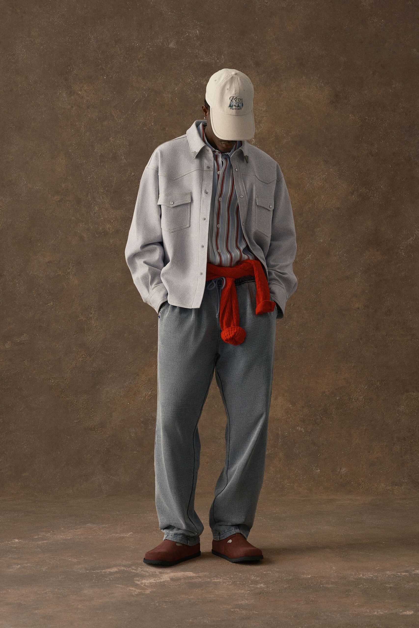 Cream suede overshirt with pearl snaps and pointed flap pockets, over a vertical striped button-down. Grey waffle-knit drawstring trousers, rust knit tied at the waist, off-white cap, brick slip-ons. Early train, platform coffee gone lukewarm.