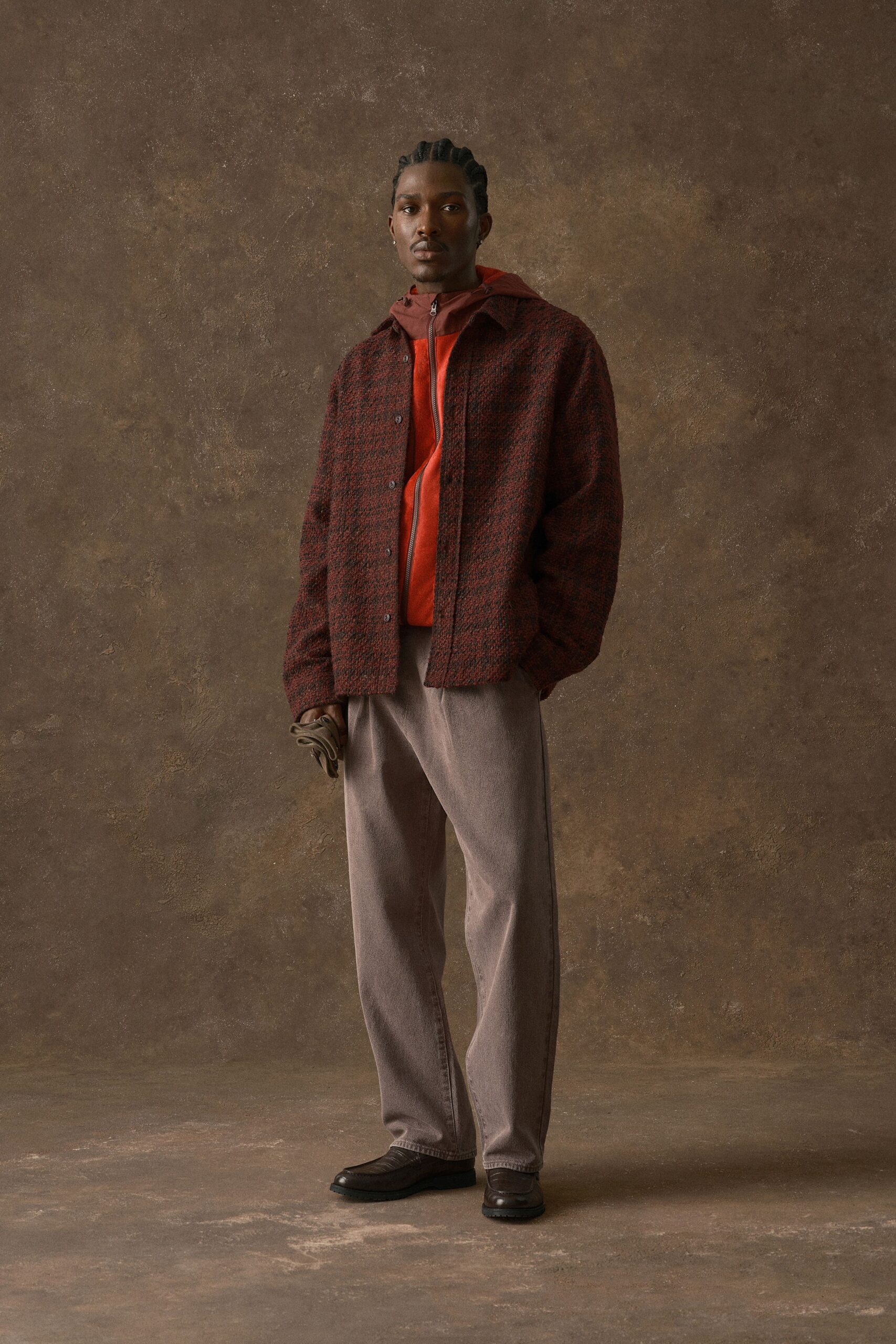Burgundy-and-black bouclé overshirt with matte snaps over a rust hooded zip jacket, the zipper pulled nearly to the chin. Taupe washed denim trousers, dark leather loafers, one glove in hand. Waiting for the train after first cold rain.