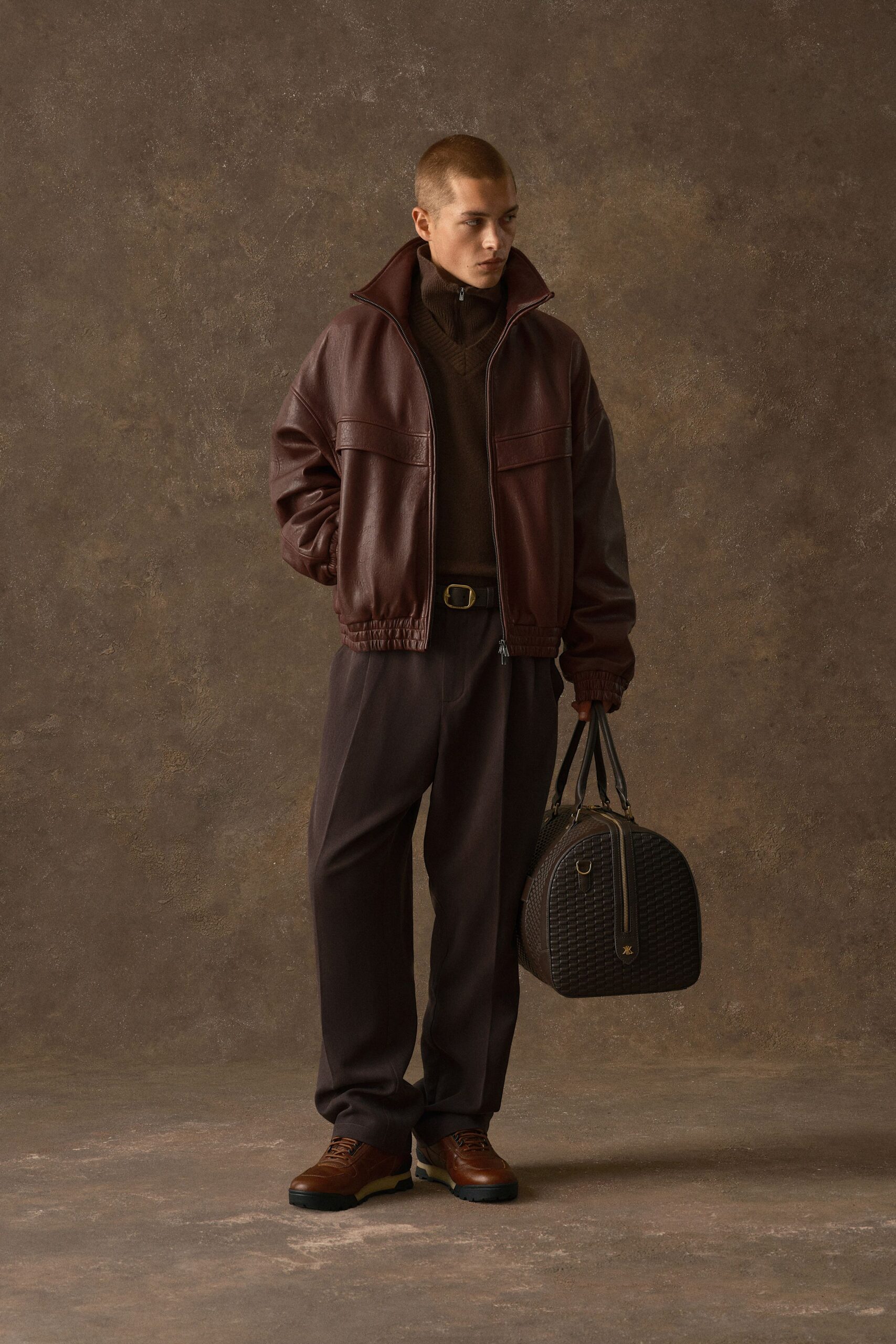 Oxblood leather blouson with a high stand collar and shirred hem, over a dark espresso ribbed zip-neck knit. Chocolate trousers with a soft front crease, tan lug-sole derbies, woven duffel. Hotel lobby before dawn.