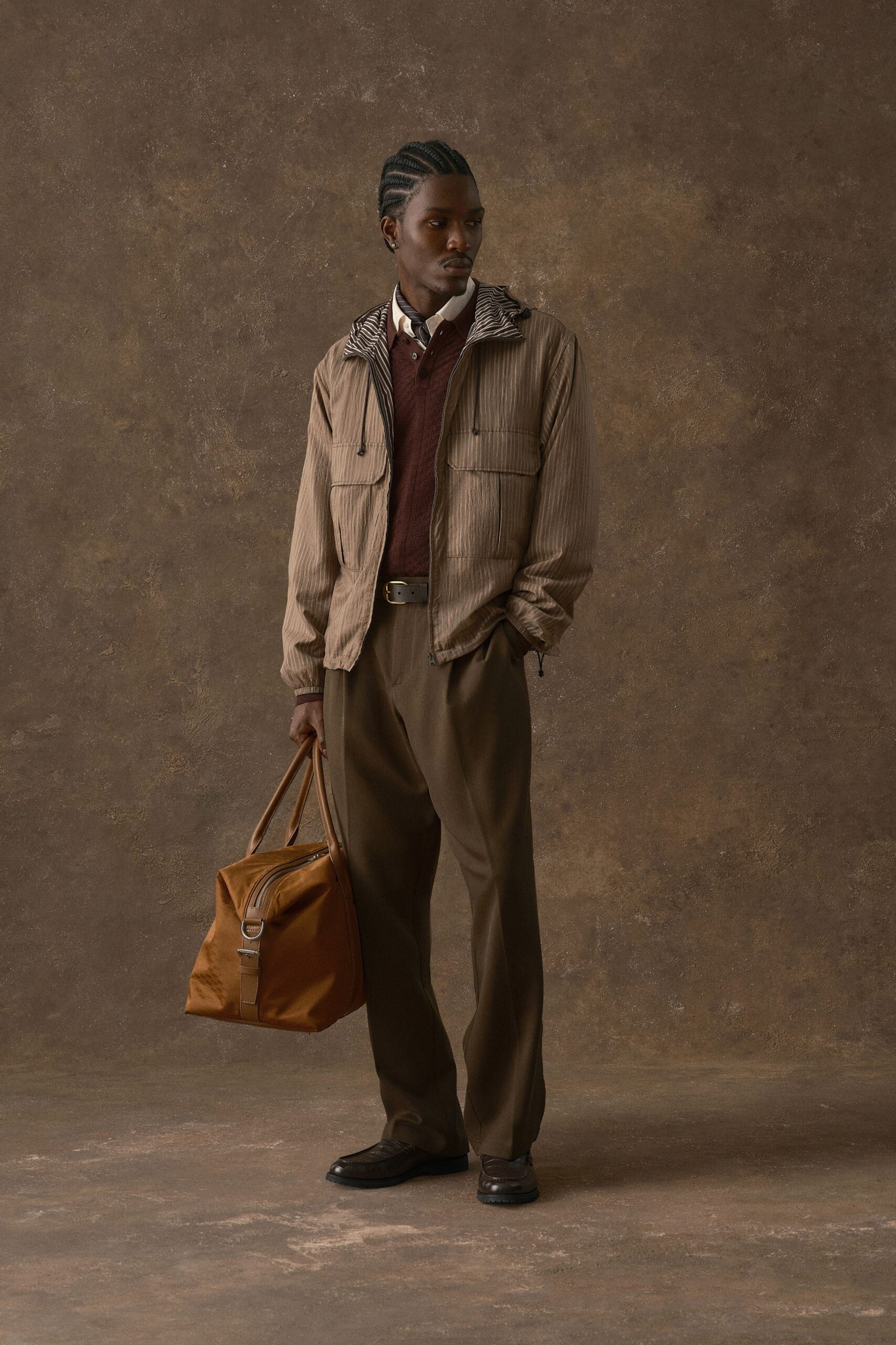 Taupe pinstriped windbreaker with zebra-striped lining at the collar, oxblood knit polo under a white point-collar shirt and narrow tie. Cocoa pleated trousers, dark loafers, tan holdall with a D-ring strap. Waiting under the station clock.