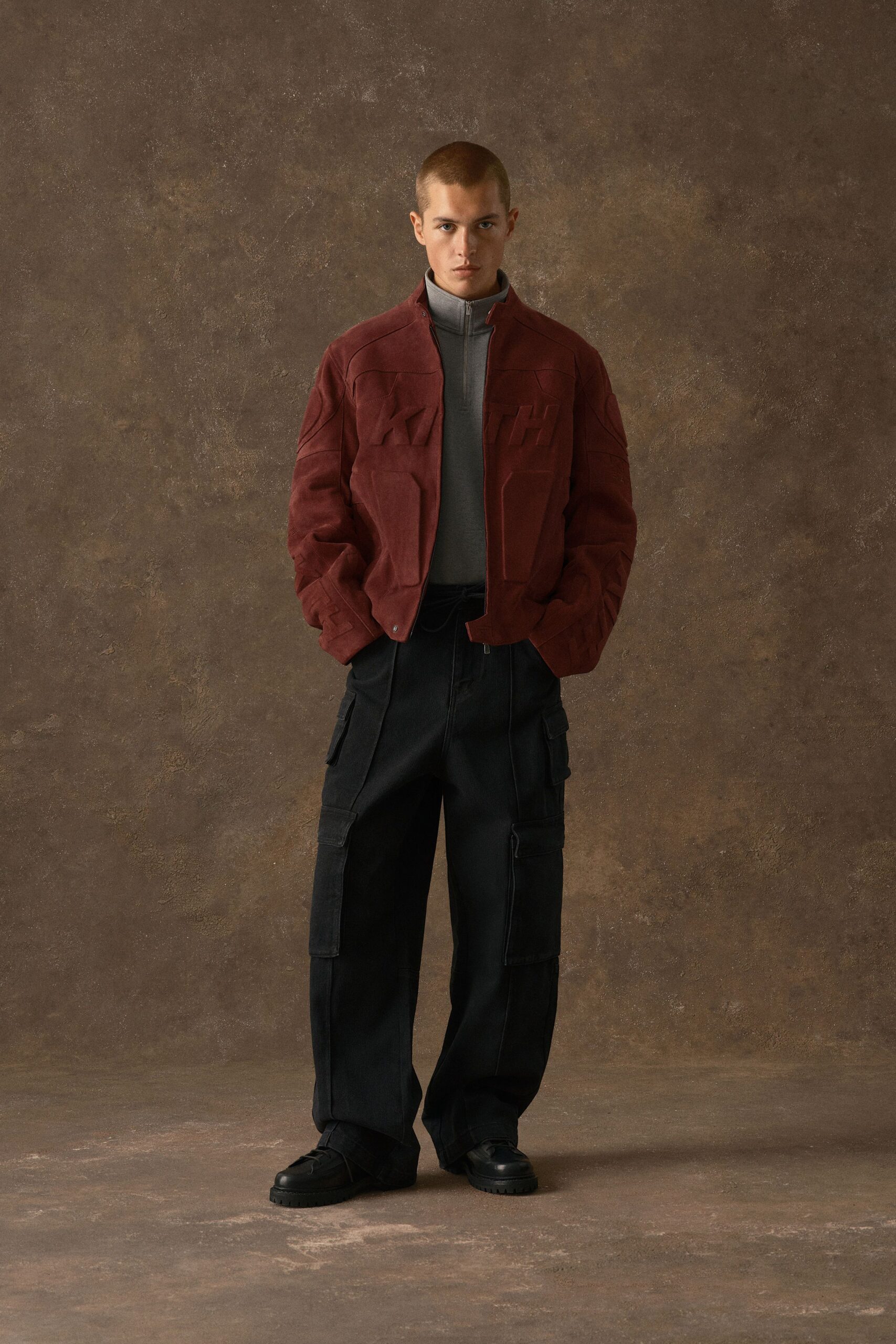 Oxblood suede bomber jacket with molded panels and a low snap tab at the hem, over a grey jersey funnel-neck zip top. Black cargo trousers with stacked flap pockets, heavy-soled shoes. Bus depot at dusk, coffee gone cold.