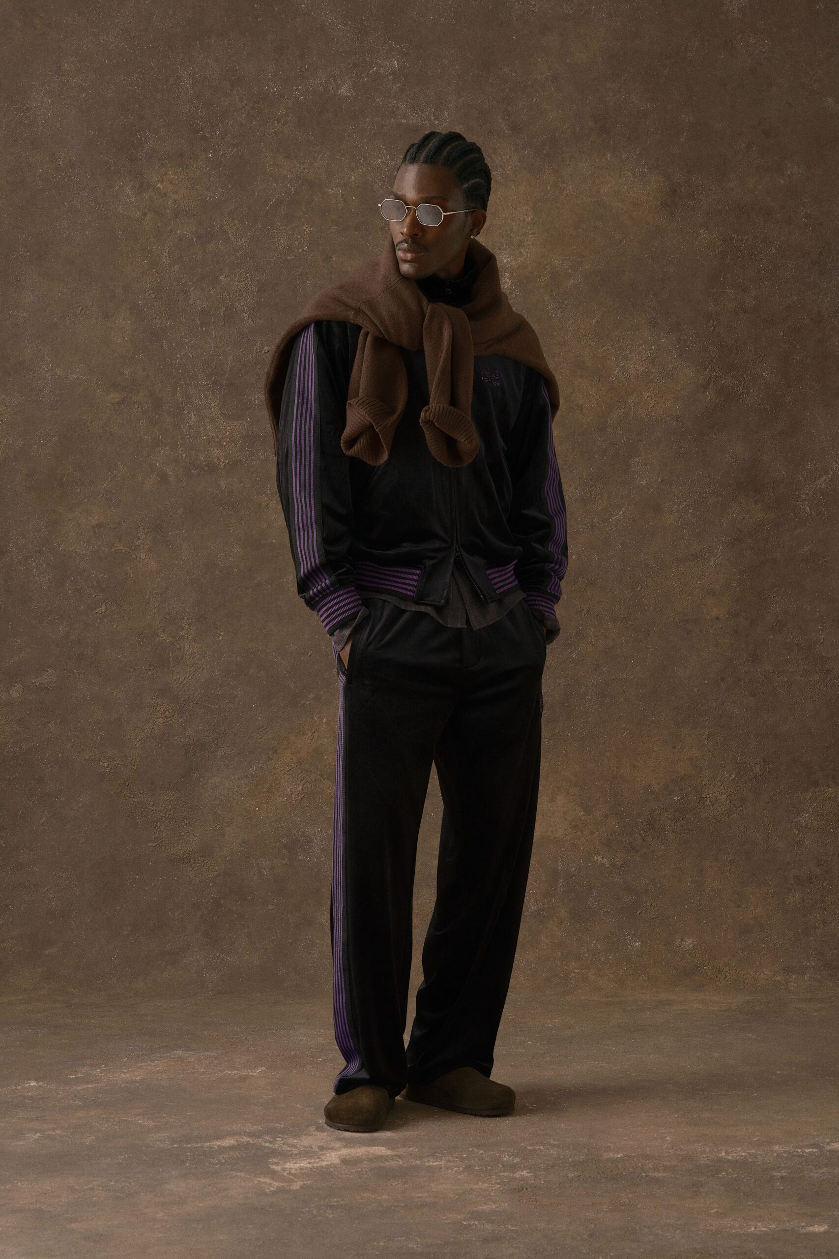 Brown wool sweater draped over the shoulders, its cuffs hanging forward. Black velour track jacket with violet rib stripes and a tiny chest motif, matching track pants, brown suede slip-ons. Hotel corridor, ice machine humming.
