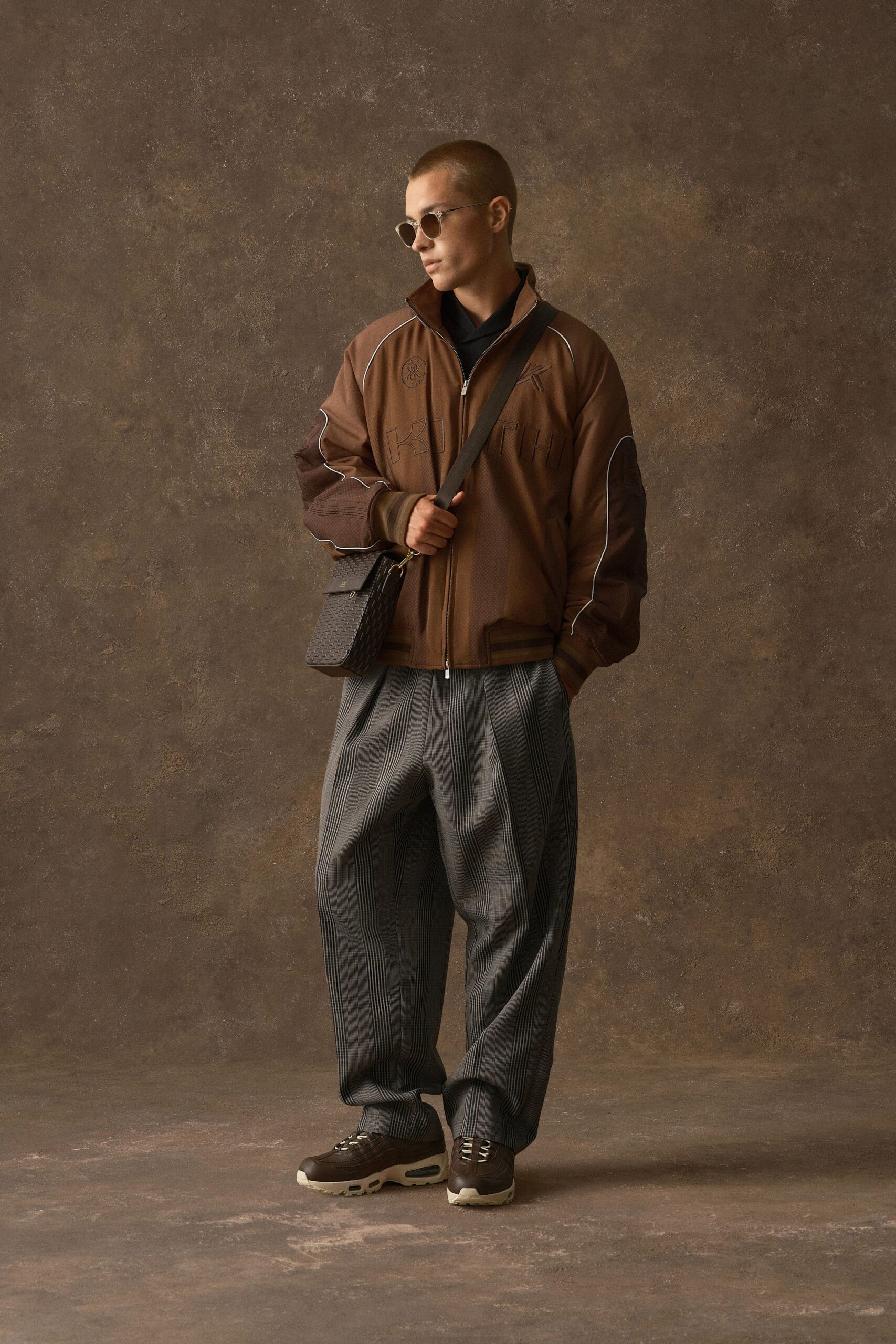 Brown zip-front track jacket with white piping tracing the sleeves, over a black collared knit. Grey glen plaid trousers with deep front pleats, brown leather trainers, woven crossbody bag. Hotel lobby, waiting for the rain to let up.