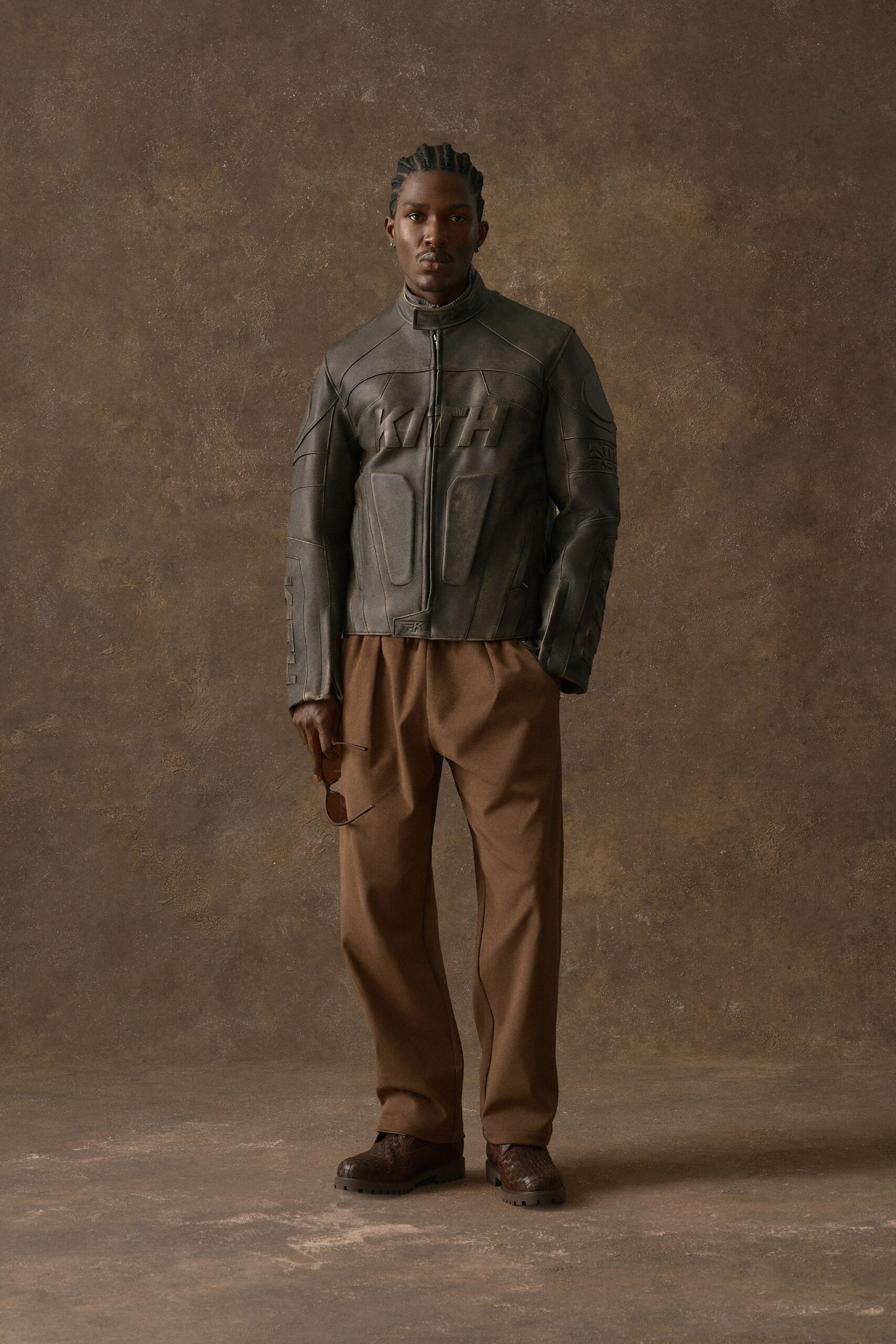 Dark olive leather moto jacket with molded panels and a stand collar zipped to the throat. Camel pleated trousers with a soft break at the shoe, brown woven leather loafers with lug soles. Waiting in a hotel lobby with dust on the ficus leaves.