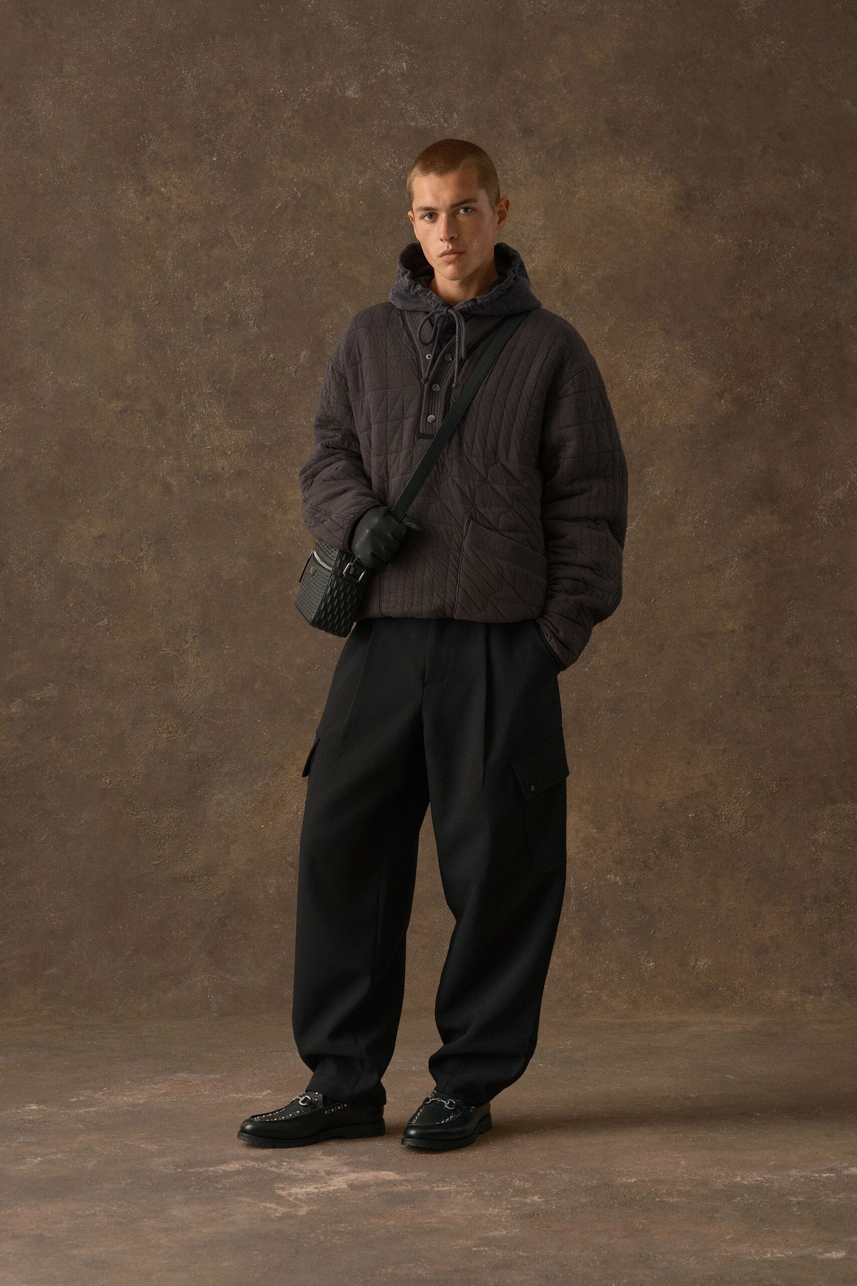 Charcoal quilted hooded pullover with a short button placket and diagonal stitch lines, black wool cargo trousers with sharp creases, smooth leather gloves, woven crossbody bag, studded loafers. Waiting on a cold station platform after dusk.