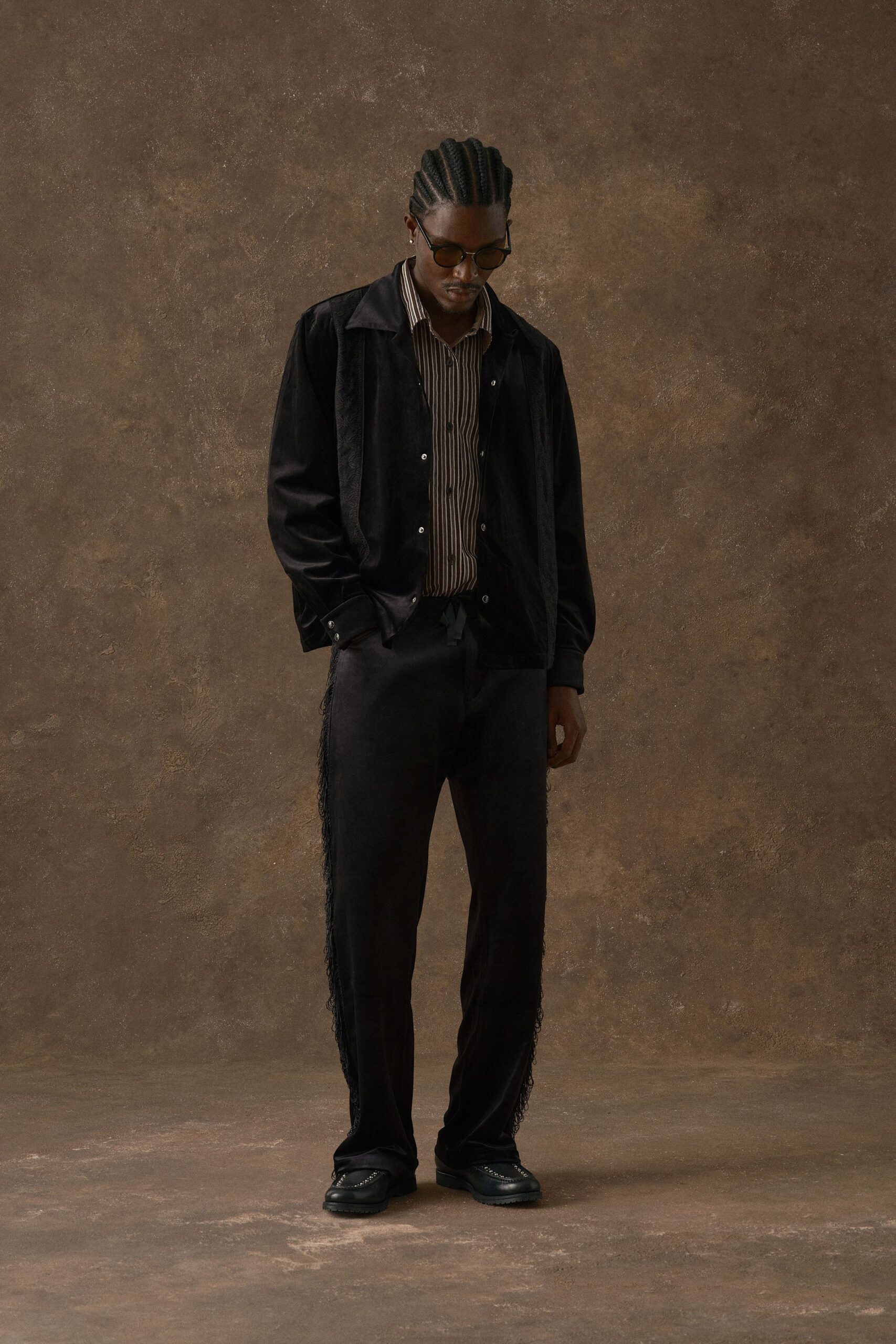 Black satin blouson with tonal jacquard panels, over a brown-and-cream striped shirt with narrow vertical pinstripes. Black trousers trimmed with long side fringe, square-toe loafers with metal studs. Waiting by the hotel bar before noon.