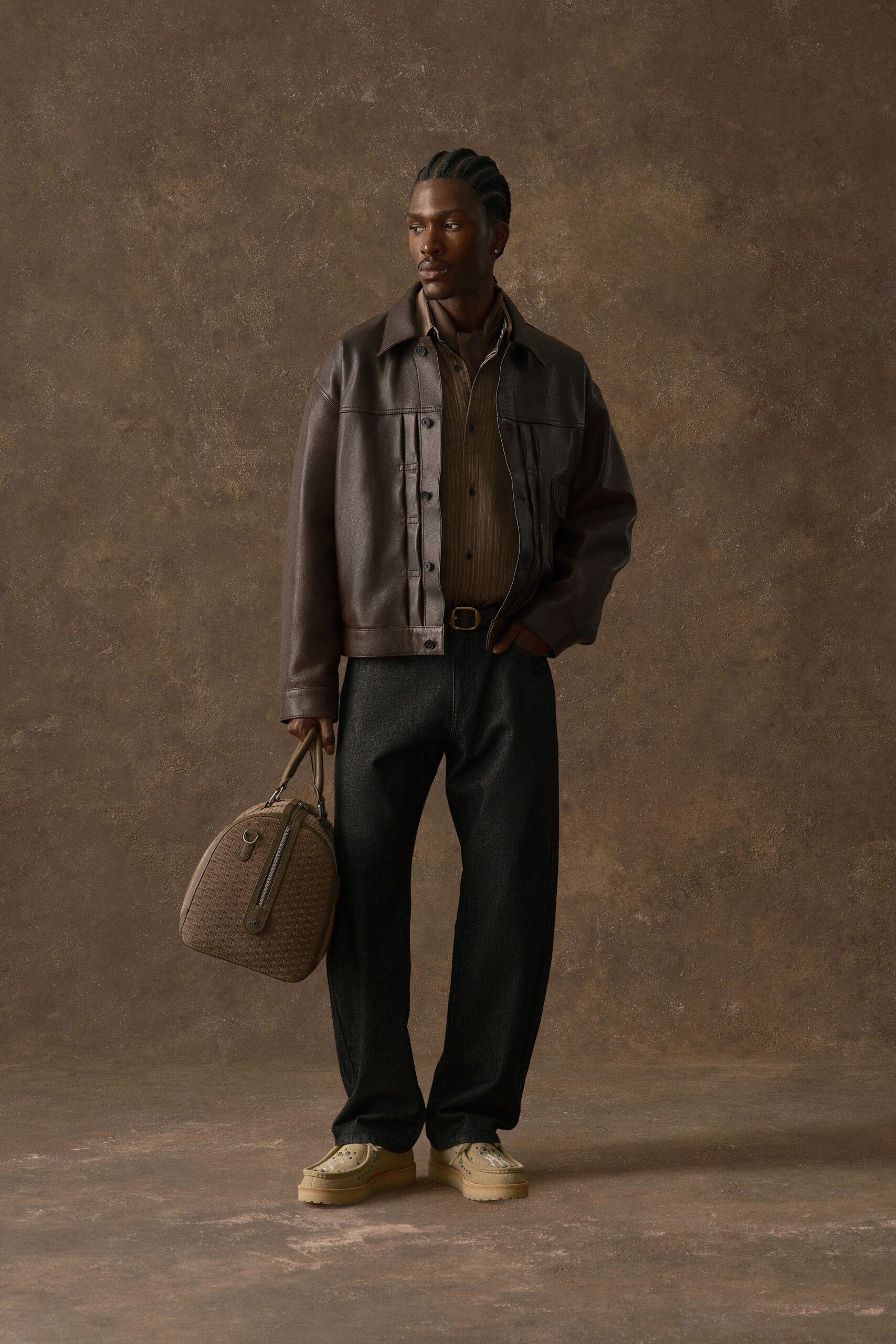 Chocolate leather jacket with a broad yoke seam, over a brown pinstriped shirt and knotted scarf. Charcoal wool trousers with a soft break, brass-buckled belt, sand moccasins studded in blue. Waiting in a hotel lobby before rain.