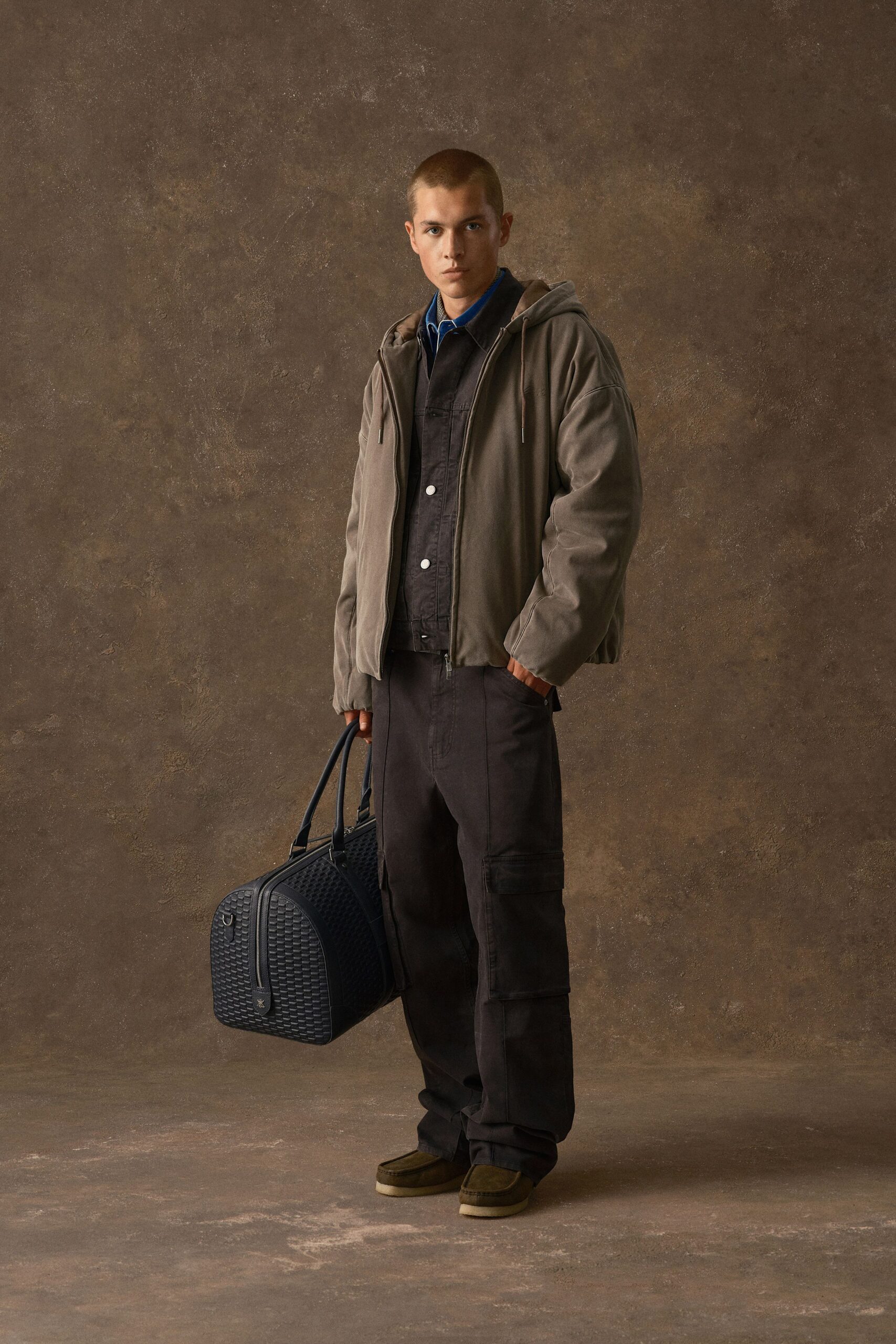 Taupe hooded jacket in brushed cotton with a two-way zip, over a charcoal denim overshirt with white metal snaps and a cobalt polo. Dark cargo trousers, suede chukka boots. Waiting by the platform heater with an overnight bag.