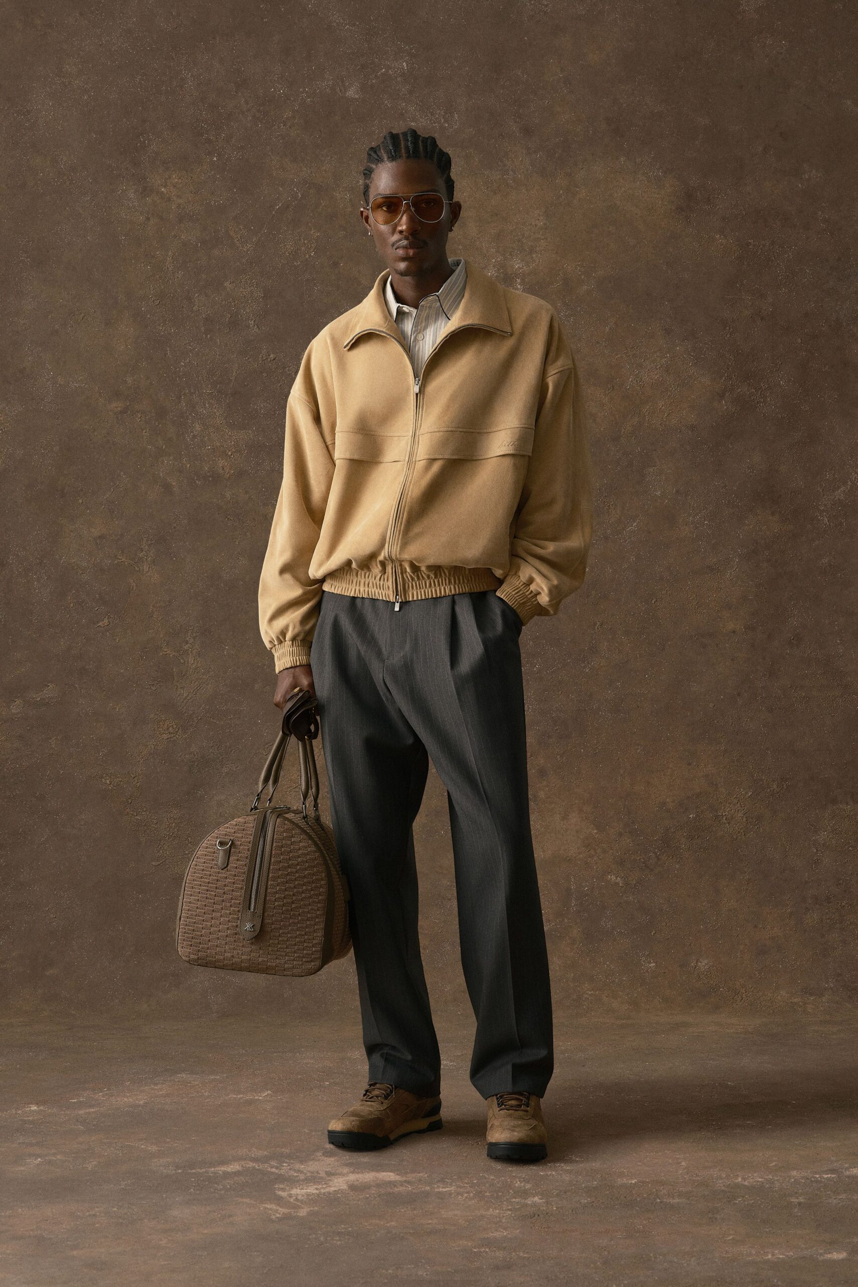 Sand suede blouson with a two-way zip and shirred hem, over a pale striped poplin shirt. Charcoal trousers fall in a soft crease to tan suede lace-ups. Brown woven holdall in hand. Early train to Milan, coffee cooling on the platform.
