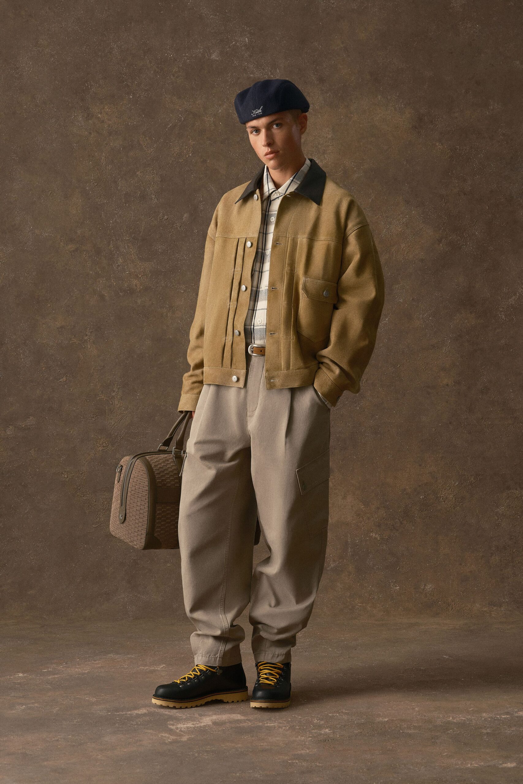 Navy wool beret, camel canvas jacket with knife pleats and a dark contrast collar, cream windowpane shirt, taupe cargo trousers with a single flap pocket, black boots laced in mustard. Waiting on a station platform with one good bag.