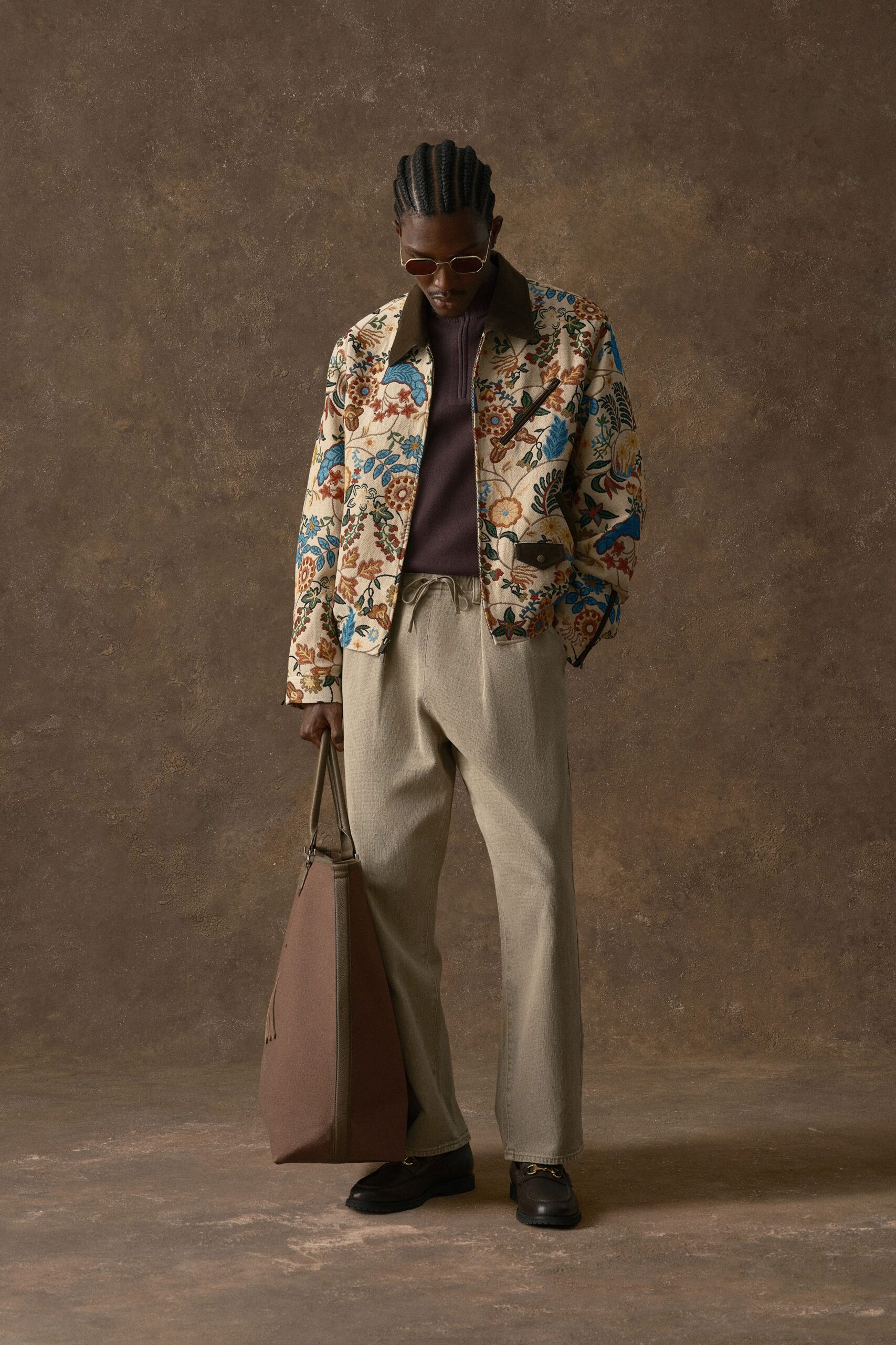 Cream floral tapestry jacket with a dark leather collar and slanted zip chest pocket, over a plum rib-knit half-zip. Oat drawstring trousers, black leather loafers, giant tan tote. Waiting out rain in a station cafe.
