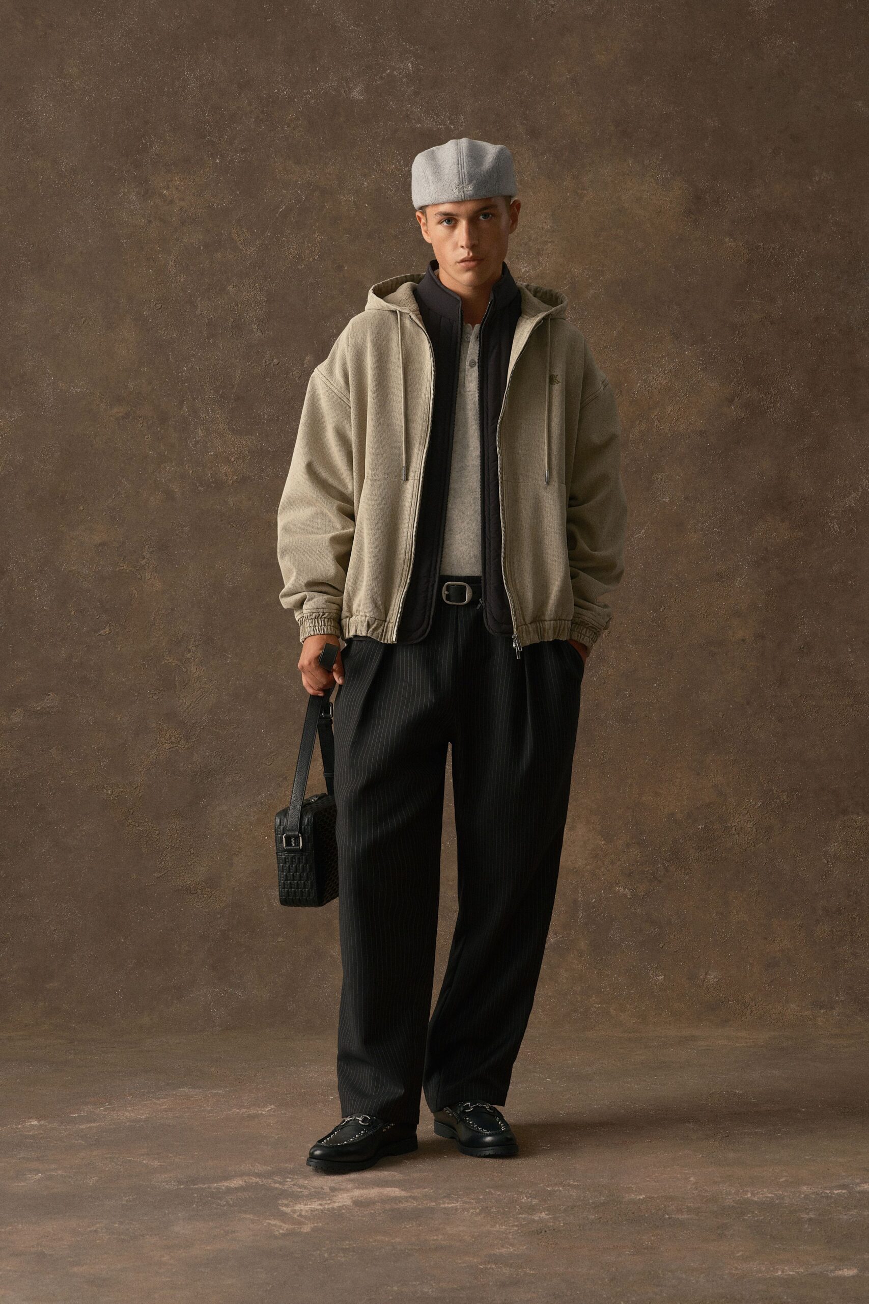 Pale stone hoodie with a cinched hem and long drawcords over a black zip-front gilet, grey heather henley, and loose black pinstripe trousers. Grey felt cap, woven leather shoulder bag, apron-toe loafers. Early train, paper cup cooling in your hand.