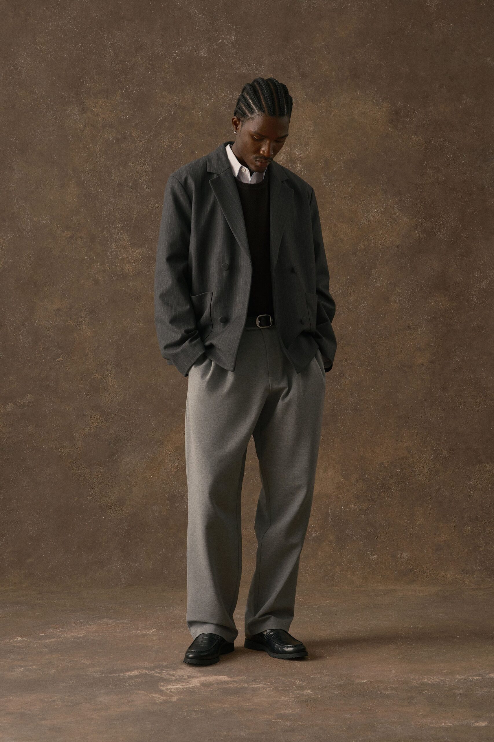 Charcoal pinstripe blazer with dropped shoulders over a white cotton shirt and black crewneck knit. Dove-grey trousers with a hard front crease, black belt with square buckle, lug-soled loafers. Waiting out a late train in the echoing station hall.