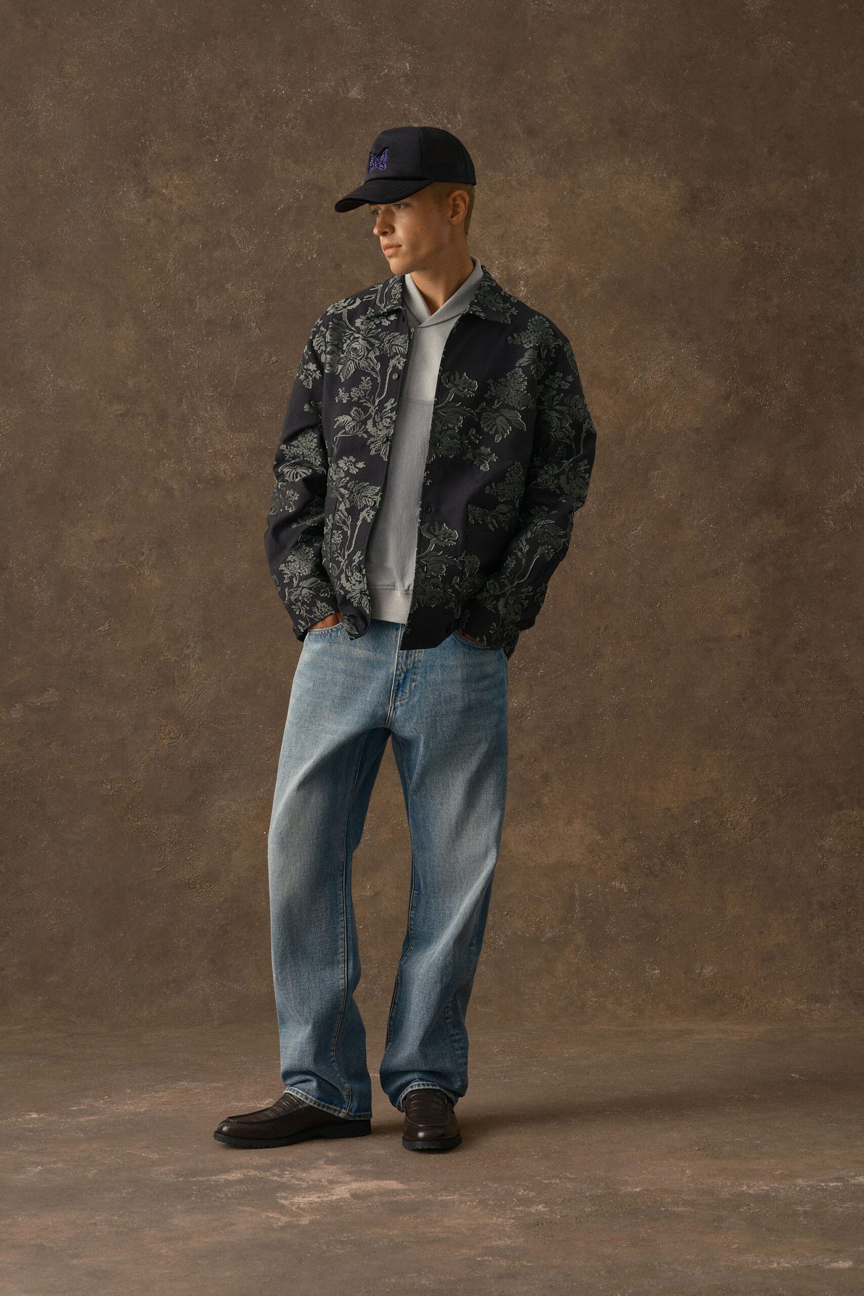 Black baseball cap with a violet embroidered butterfly, dark jacket woven with smoky floral jacquard, pale grey funnel-neck knit, light-wash jeans with a soft whisker fade, brown moc-toe loafers. Waiting by the freight elevator after soundcheck.