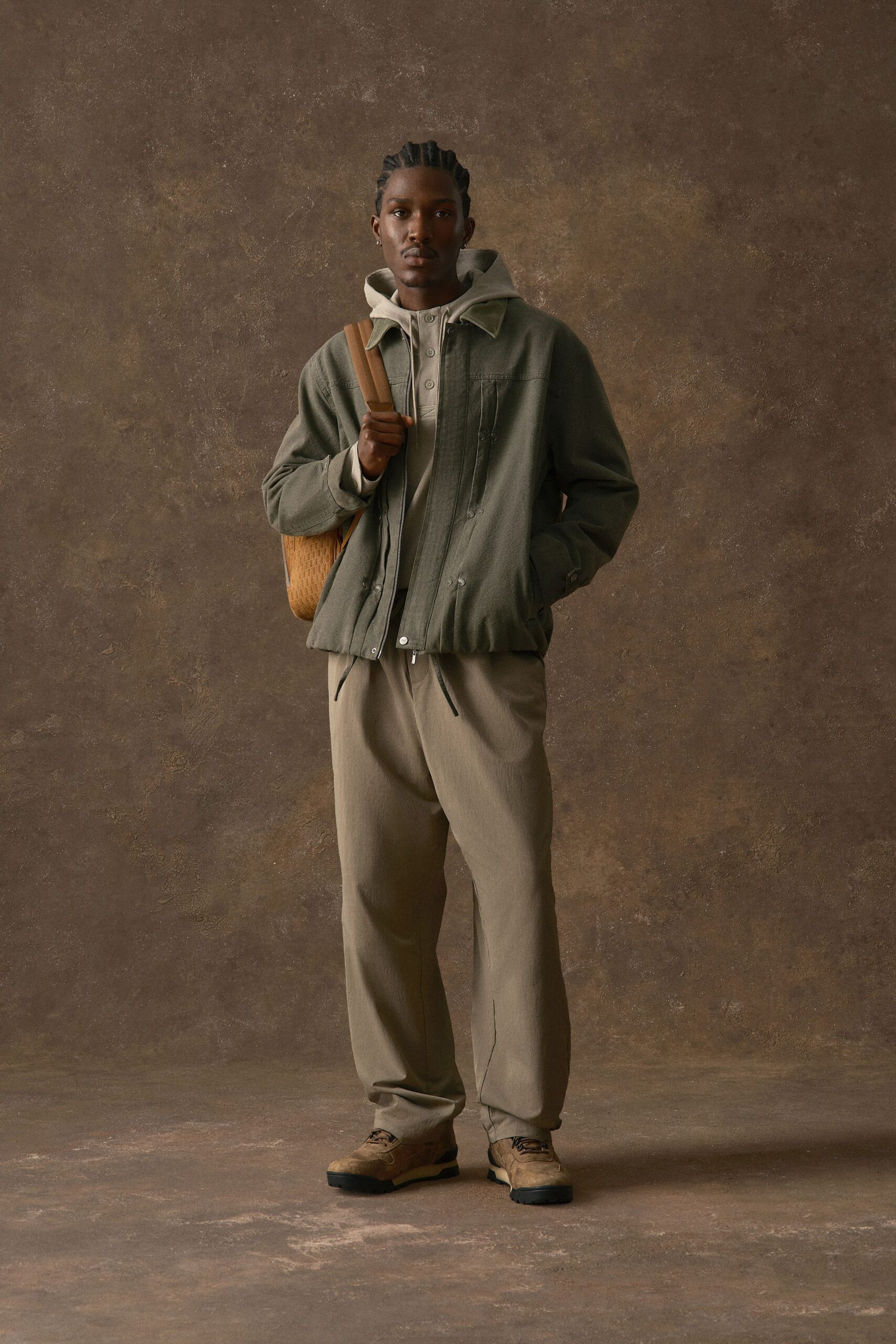 Olive canvas jacket with a zip front and puffed drawcord hem over a stone waffle-knit henley hoodie. Taupe trousers falling loose to suede derby shoes. Woven tan shoulder bag tucked under one arm. Early train, platform grit underfoot.