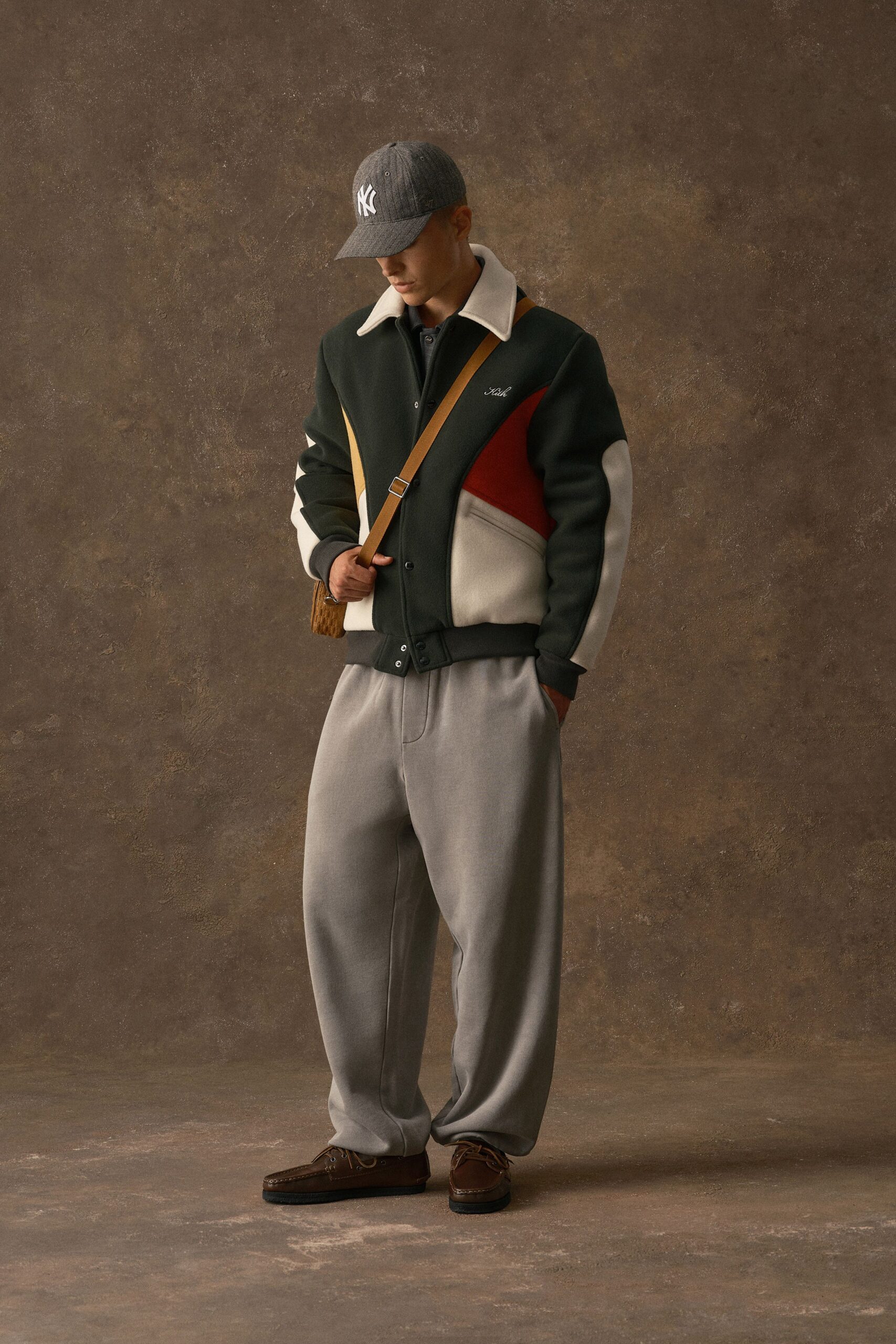 Grey pinstripe baseball cap, dark wool bomber with a cream felt collar and rust colour-block panels, charcoal polo beneath. Dove-grey trousers with a pressed front crease, brown moccasins, tan strap bag. Hotel lobby at 7am, waiting on coffee.