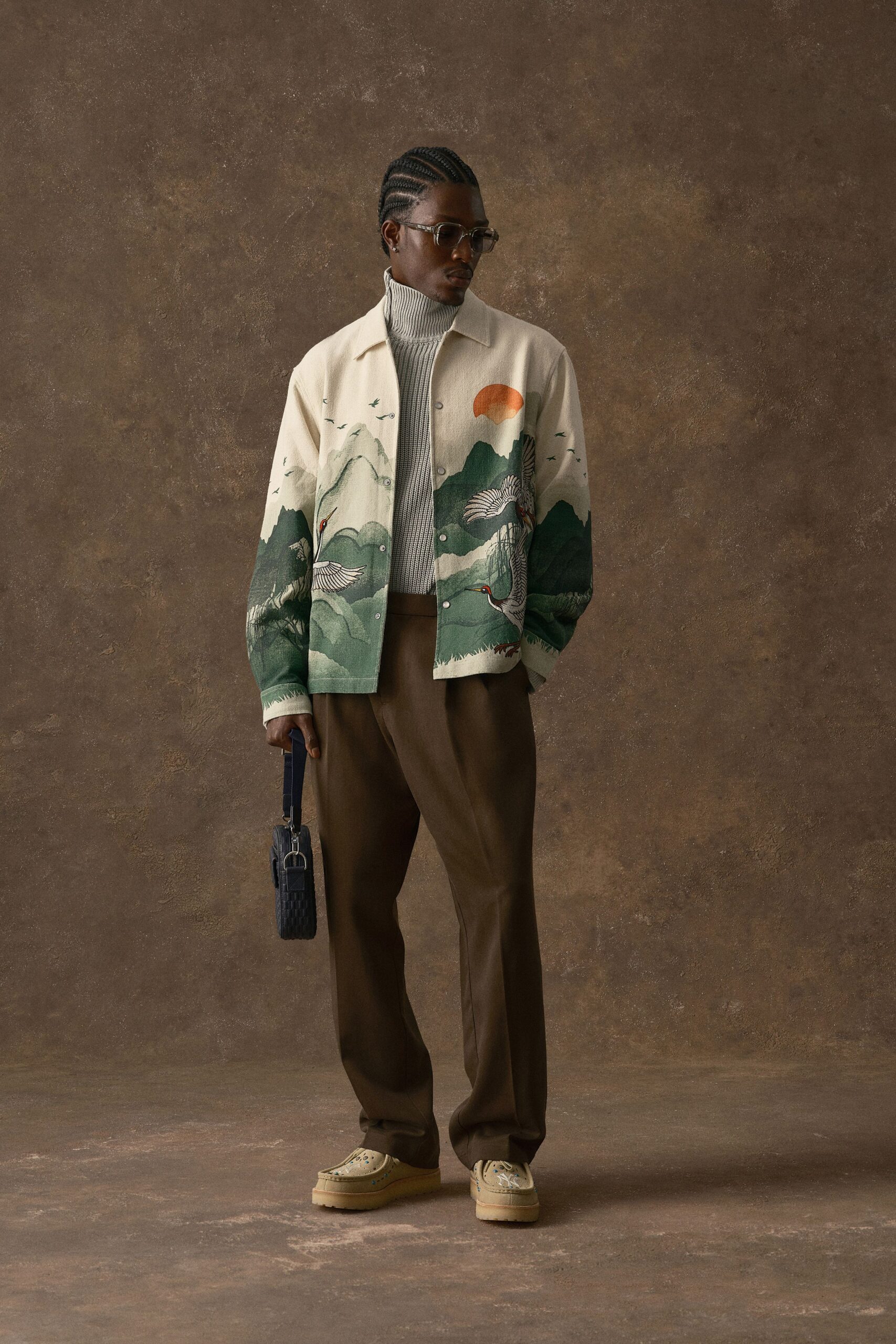 Ivory overshirt printed with cranes and pine-dark mountains, fastened with matte snap studs over a greige rib-knit turtleneck. Espresso pleated trousers, cream clogs with punched motifs. Waiting out a delayed train near the tea kiosk.