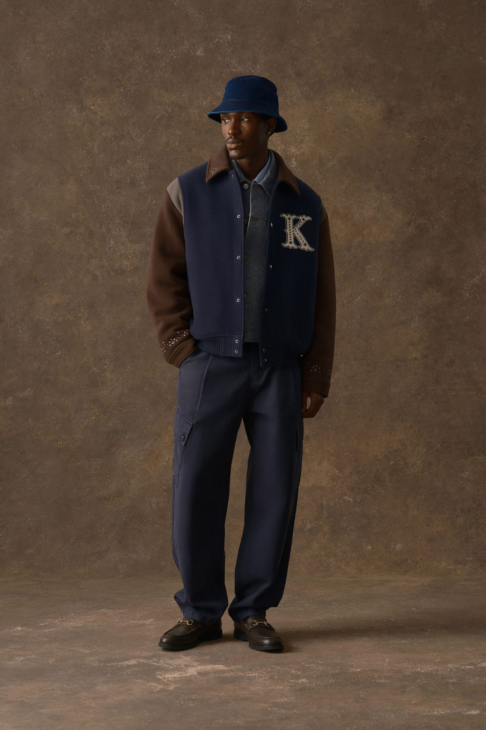 Navy bucket hat in brushed felt, varsity jacket with brown sleeves and tiny studs circling the cuffs, slate knit polo, loose navy cargo trousers with flap pockets, dark loafers with horsebit hardware. Waiting in a hotel bar before noon.
